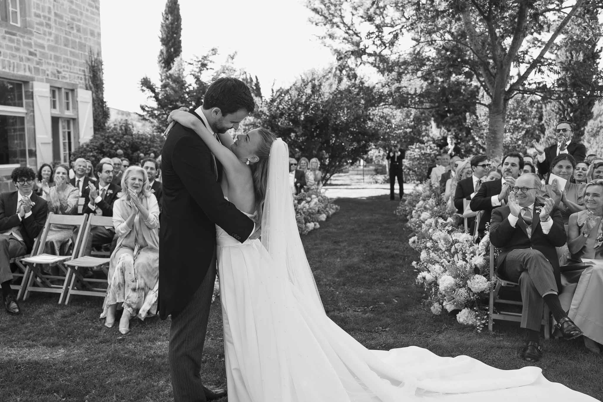Black and white first kiss moment with 50 guests applauding at outdoor garden ceremony