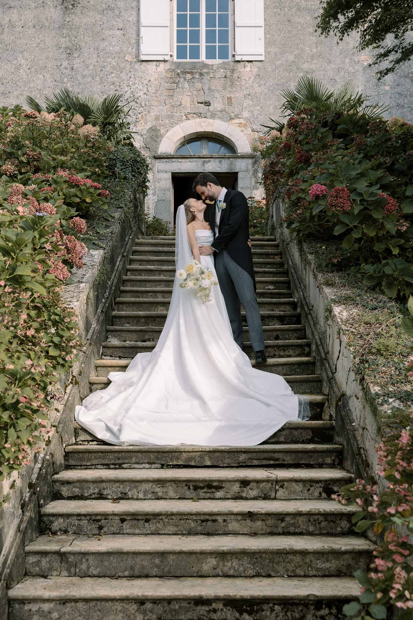 Couple kisses on chateau stone staircase with cathedral train cascading down steps and pink hydrangeas