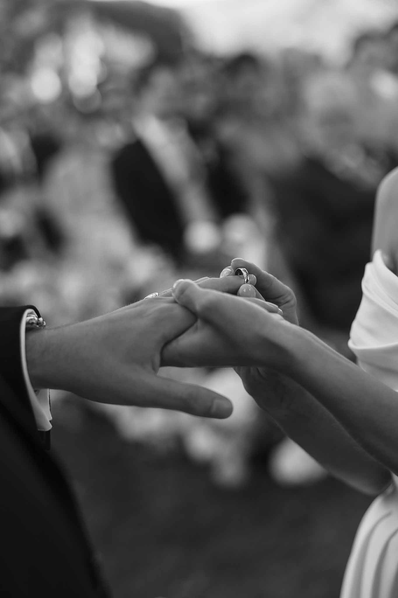 Close-up of ring exchange moment with groom sliding band onto bride's finger with bokeh guests behind in B&W