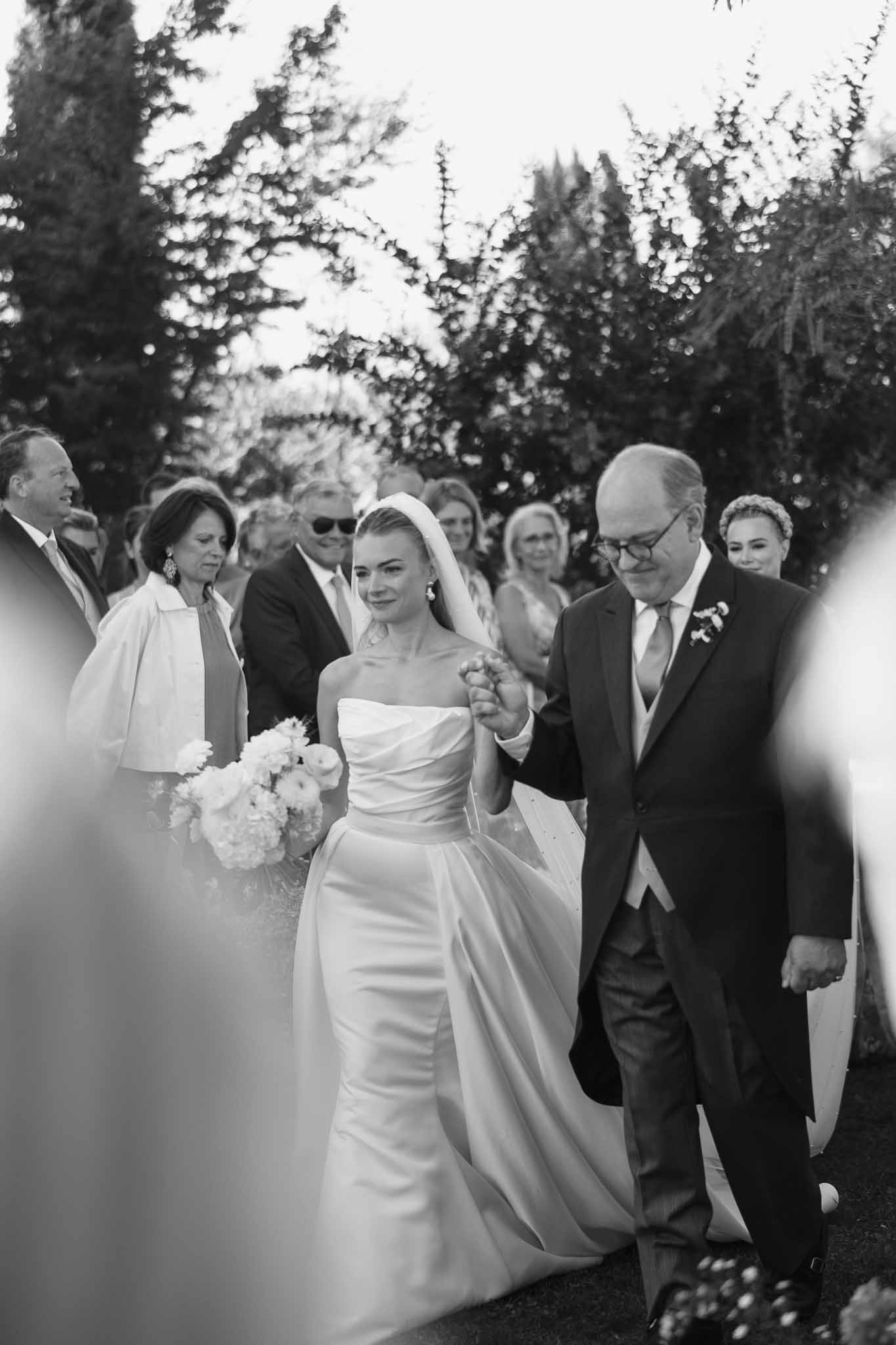 Black and white candid of father walking bride down outdoor aisle with hydrangea bouquet and veil