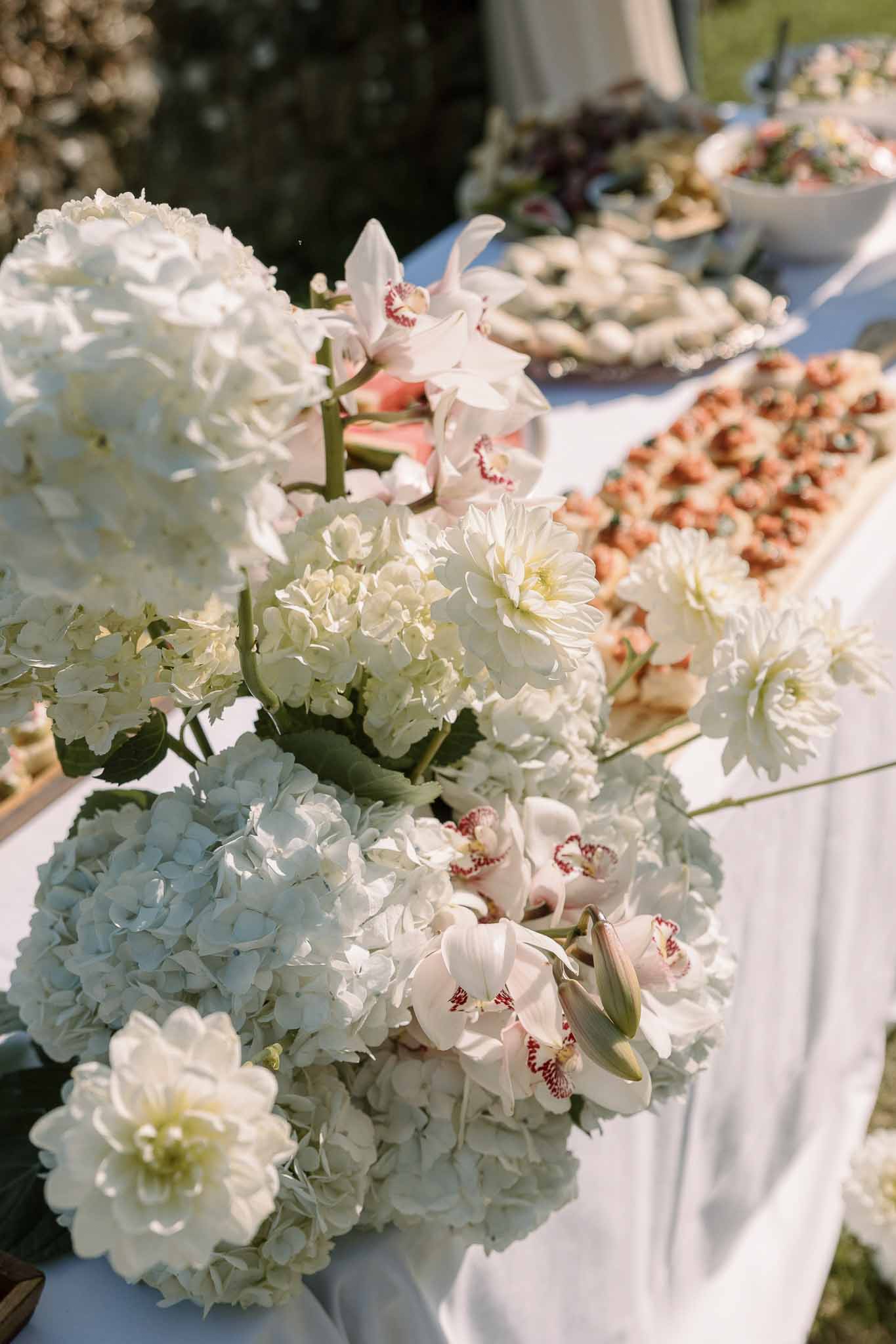 Cocktail hour catering table with white hydrangea and orchid arrangement alongside canape platters outdoors