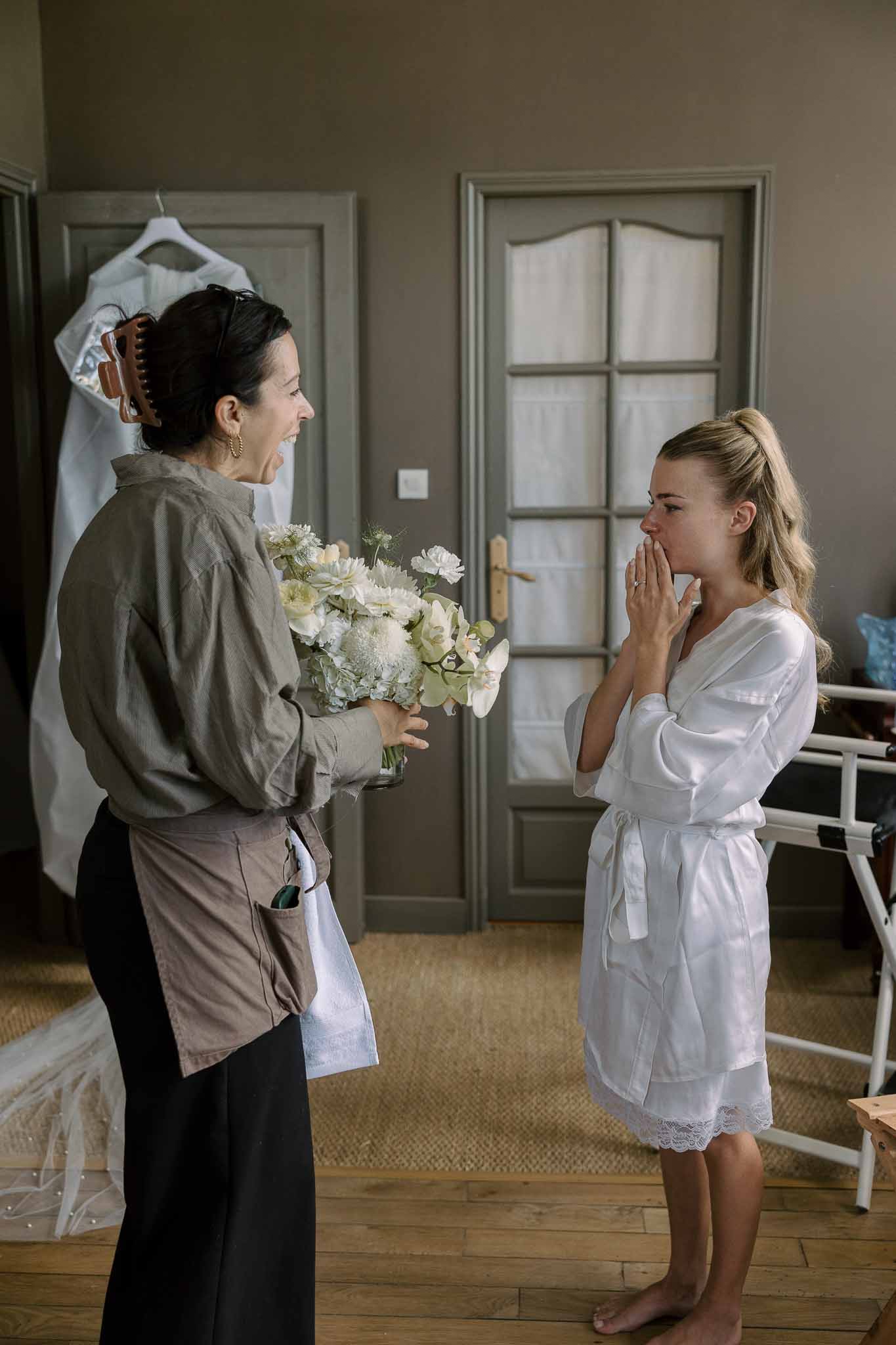 Bride in white robe reacting emotionally as florist presents white and green bridal bouquet
