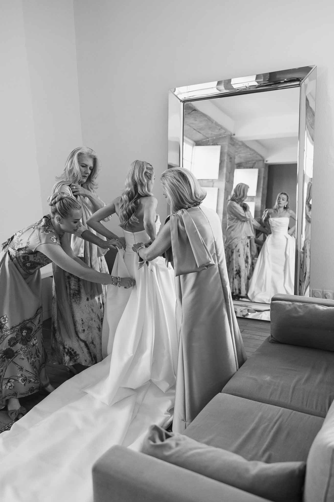 Three women help bride into strapless ballgown with mirror reflection in modern suite interior in B&W
