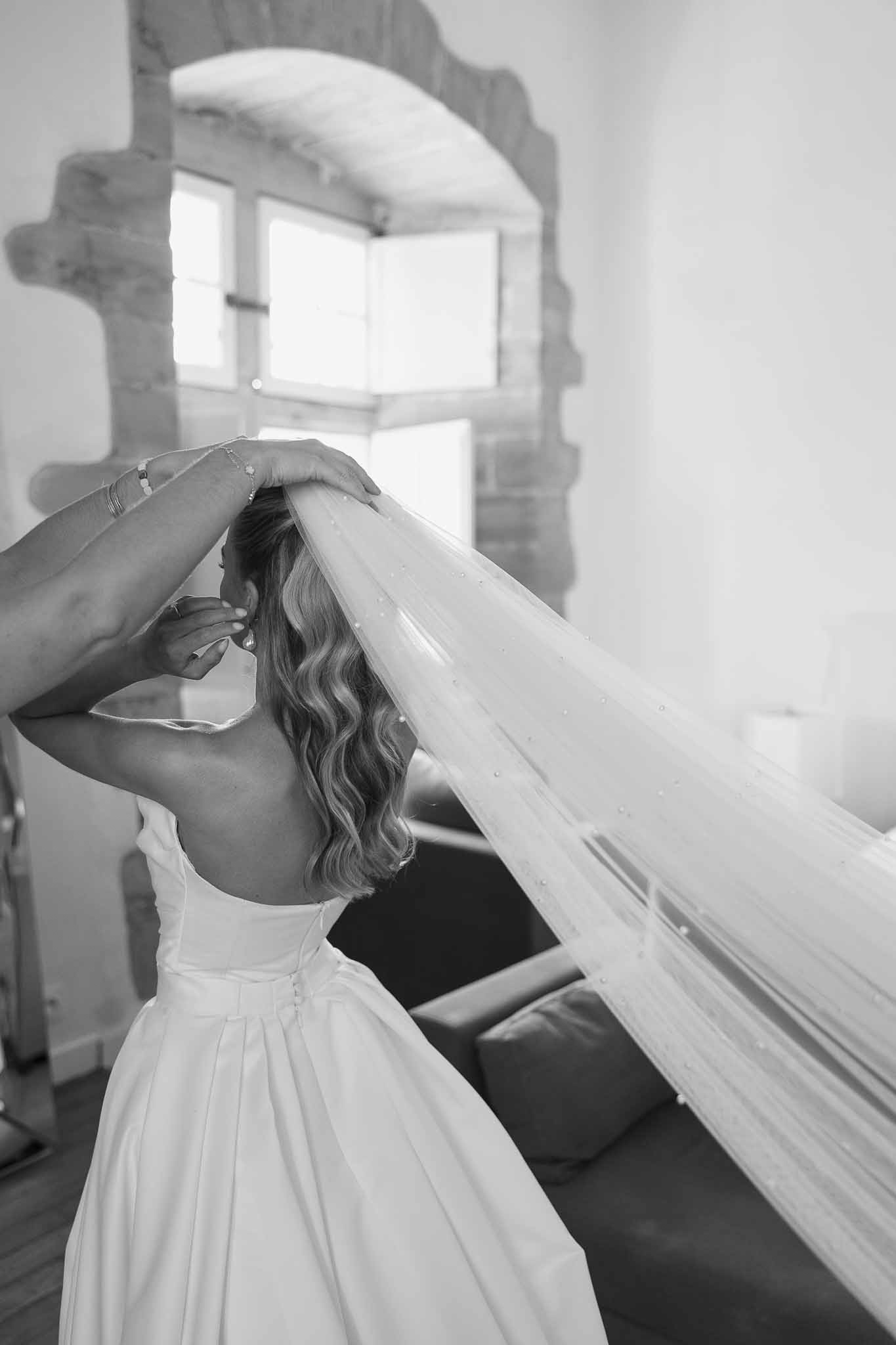 Black and white portrait of bride from behind placing a pearl-edged veil into her hair near a rustic mirror