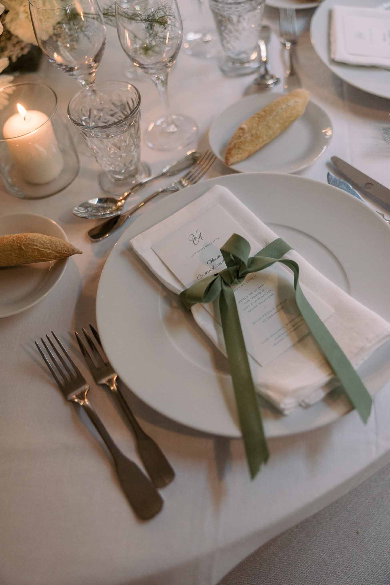 Place setting with ivory napkin tied in sage ribbon, calligraphy escort card, crystal glasses, and pillar candle
