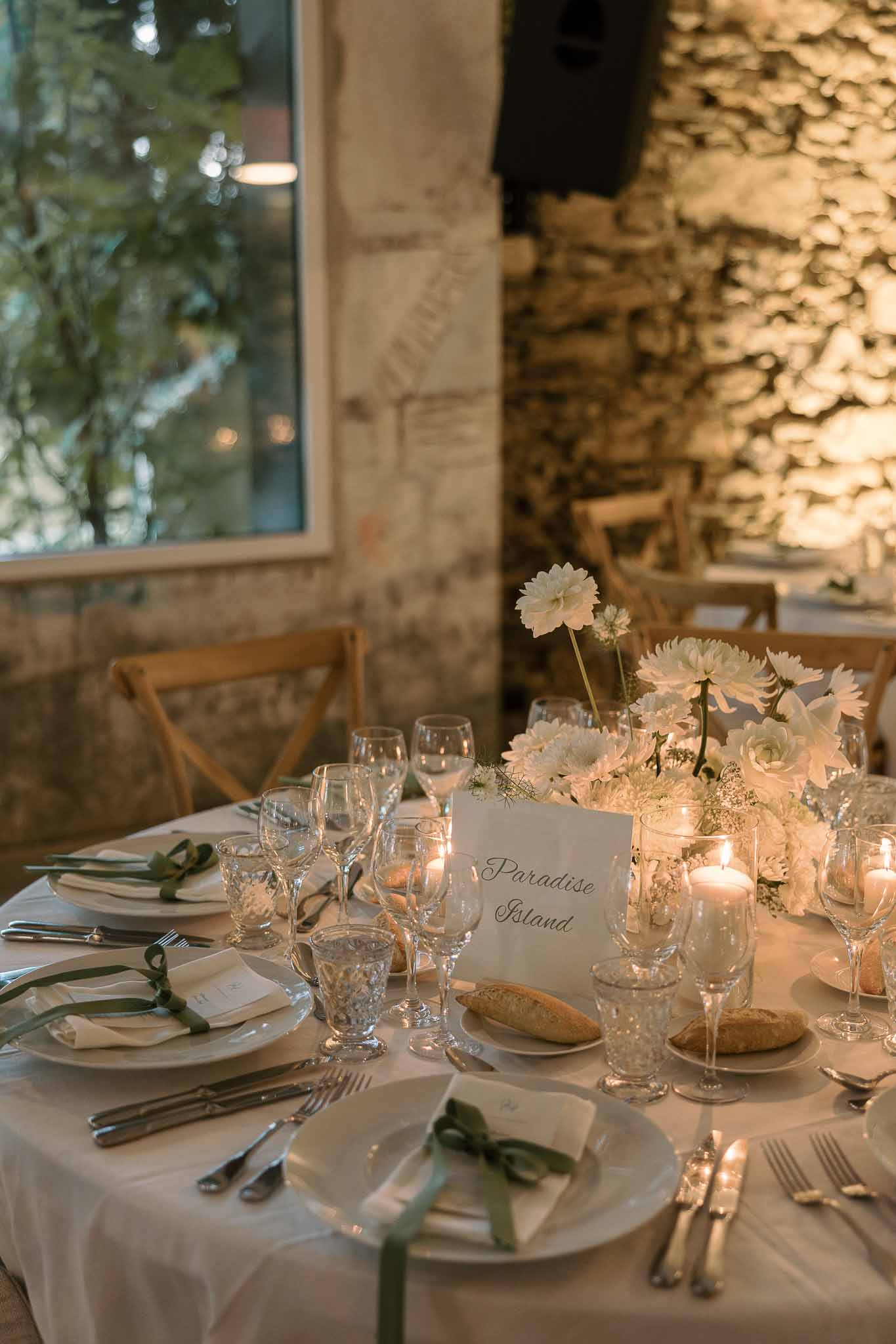 White dahlia centerpiece with sage ribbon napkins and calligraphy table card beside crystal glassware on stone wall