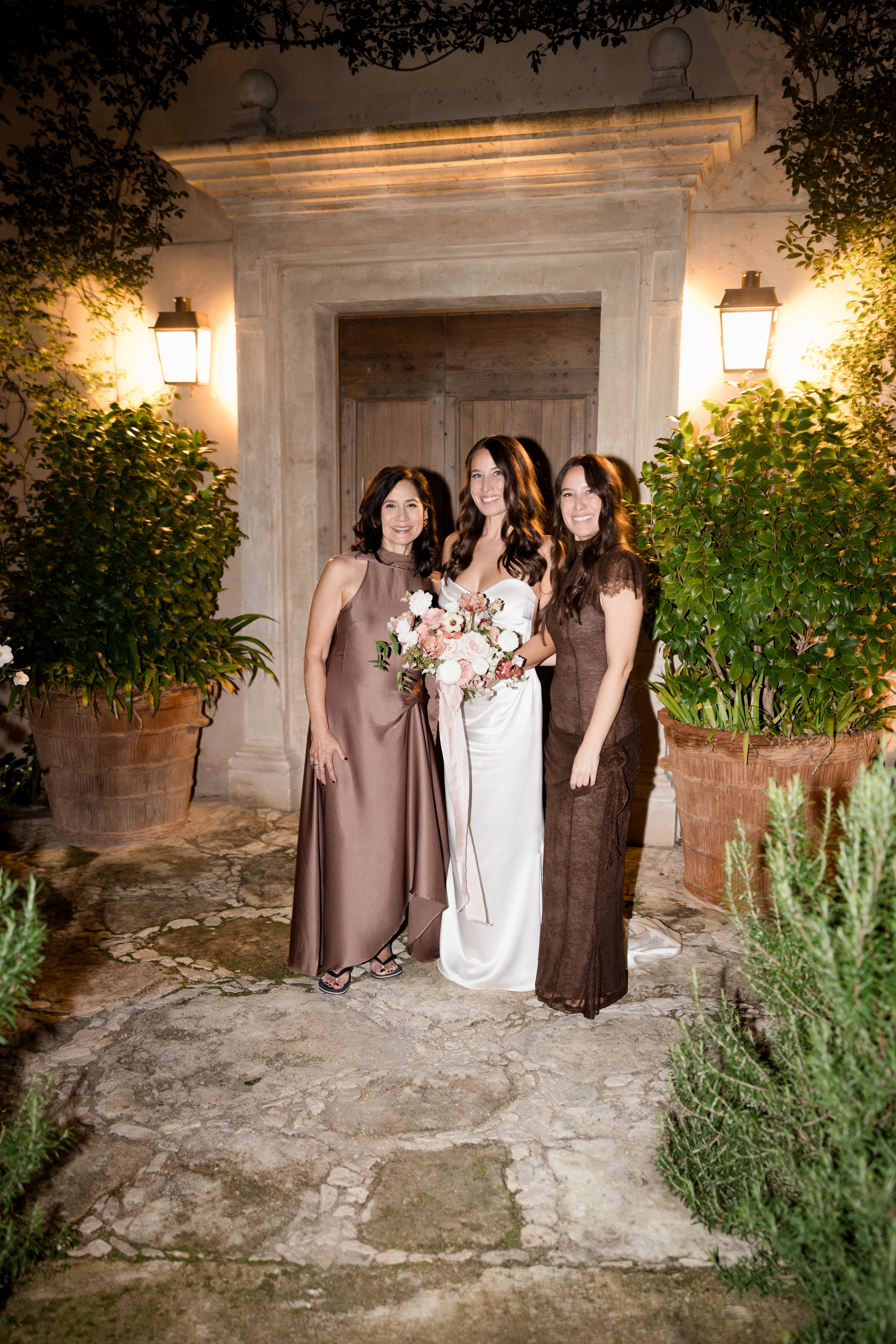 Bride in ivory satin gown with two attendants in brown dresses posing at night in front of stone chateau doorway