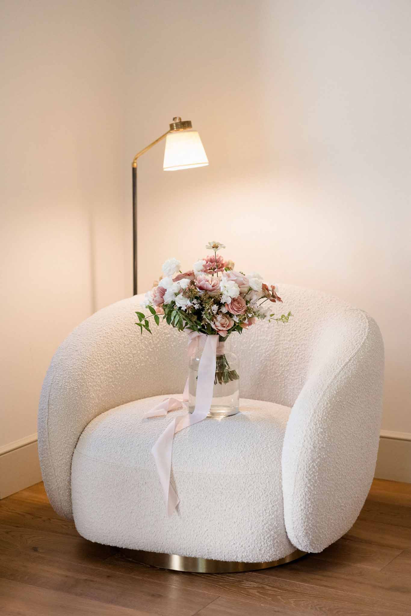 Bridal bouquet of dusty rose roses and blush peonies with trailing satin ribbons on a cream boucle armchair