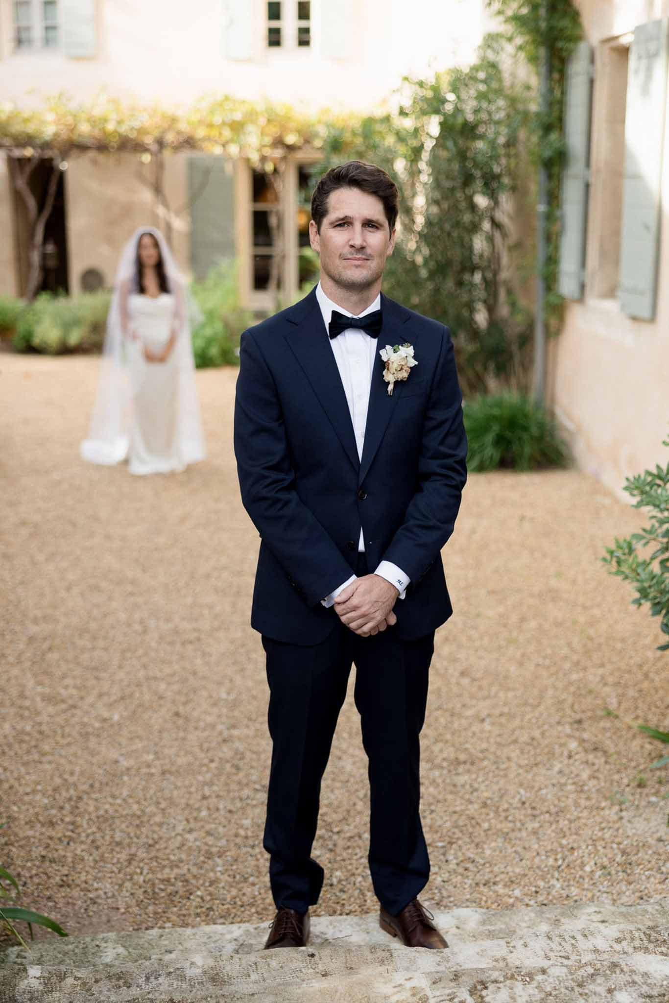 First look with groom in navy tuxedo waiting as bride approaches in soft focus at Provencal courtyard