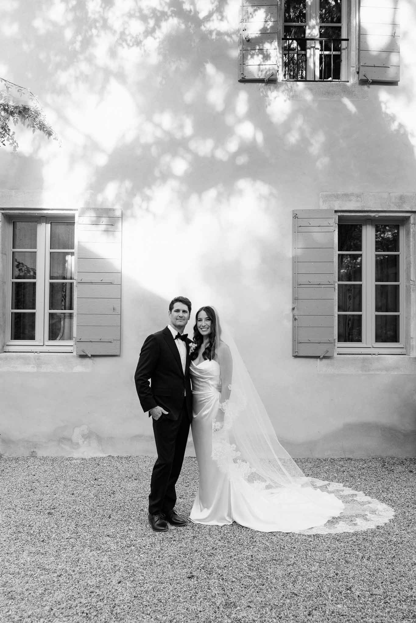 Bride in satin cowl gown with lace-edged cathedral veil and groom in tuxedo before country house in B&W