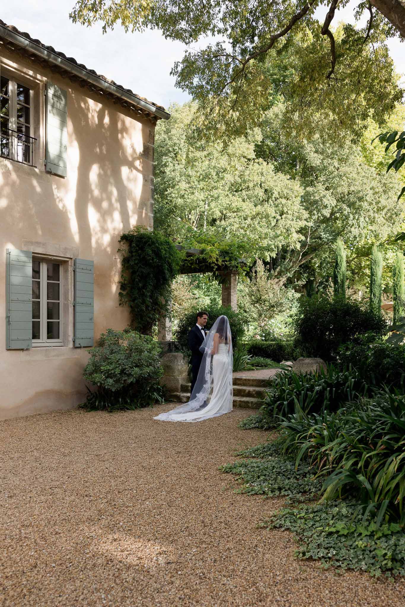 Couple facing each other beside Provencal mas with peach walls, sage shutters, and trailing lace veil