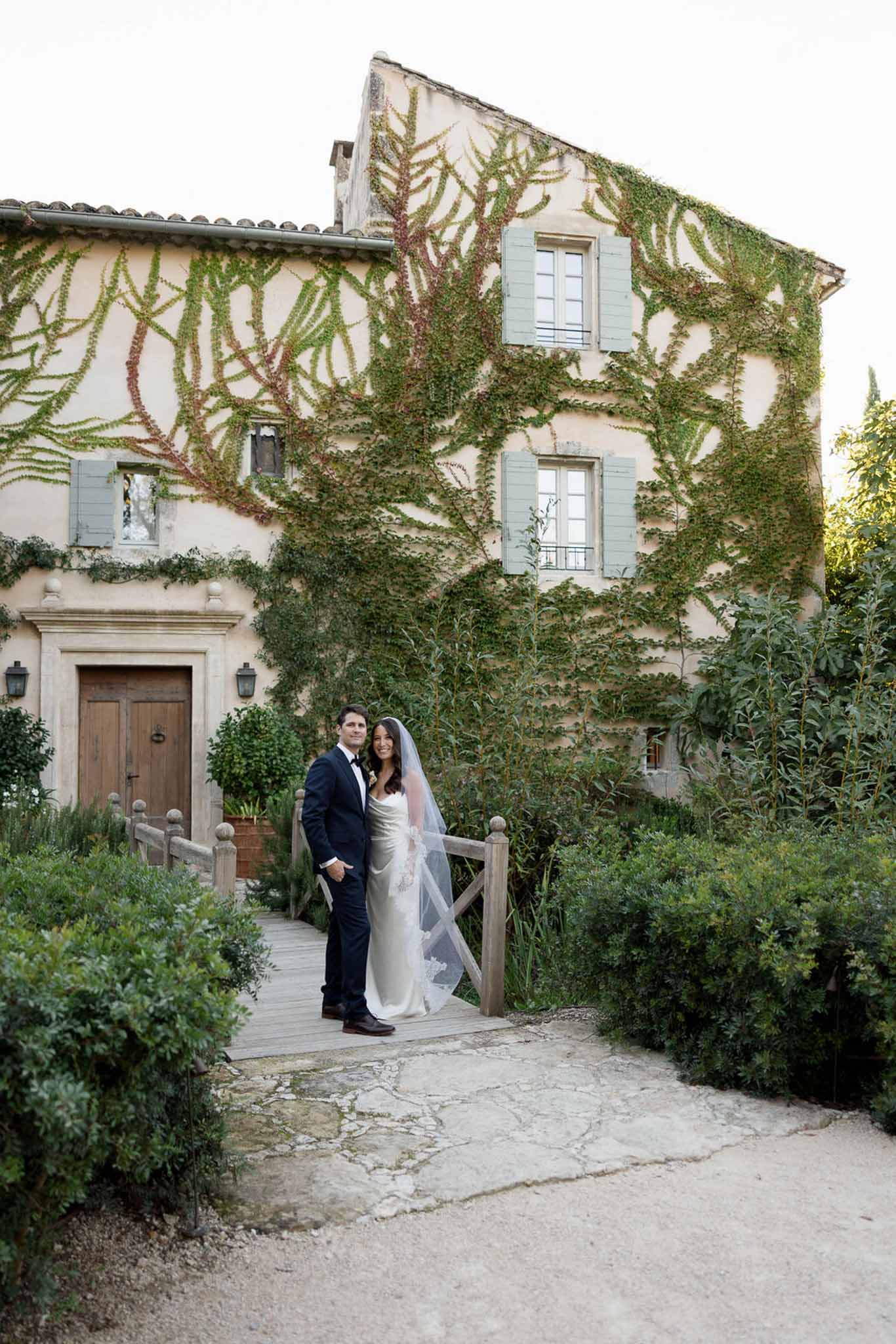 Couple on wooden bridge before ochre mas with sage shutters and ivy, bride in lace-edged veil