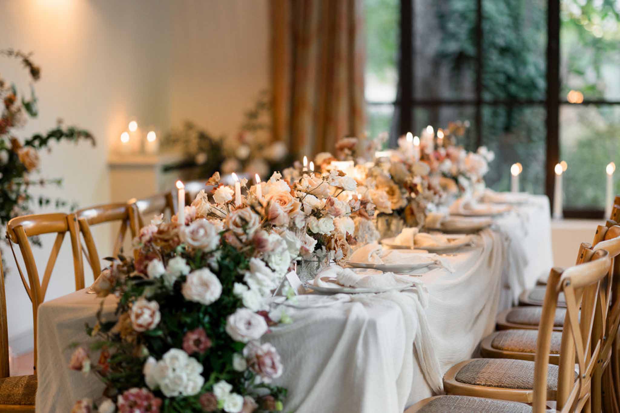 Head table with blush, peach, and dusty mauve rose runner and white taper candles before fireplace mantel indoors