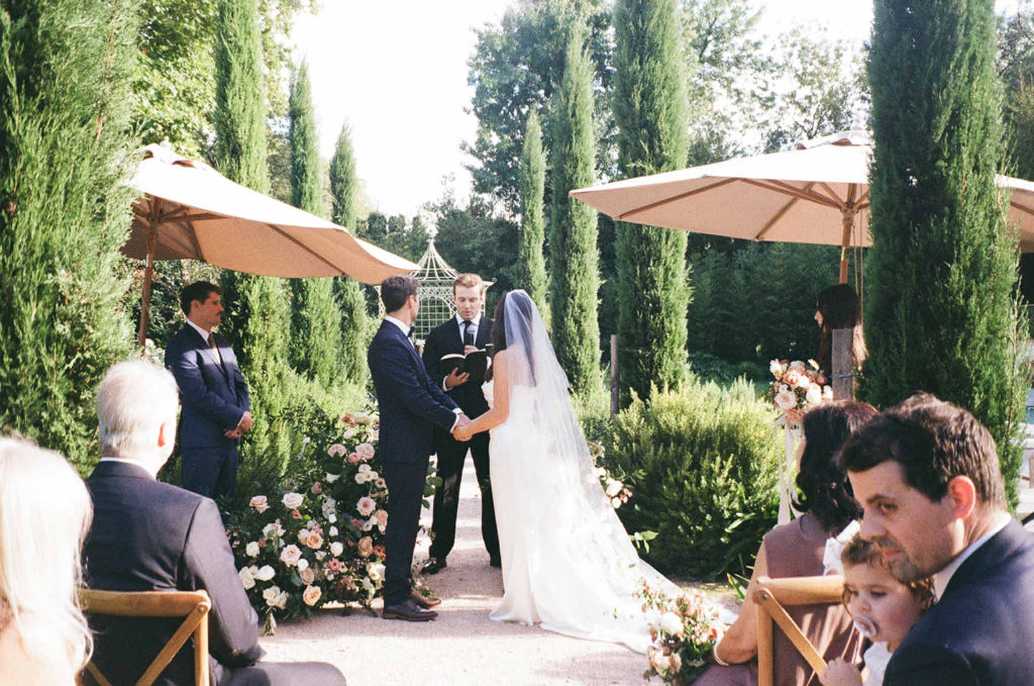 Outdoor wedding ceremony between cypress trees with bride in cathedral veil and blush rose aisle arrangements