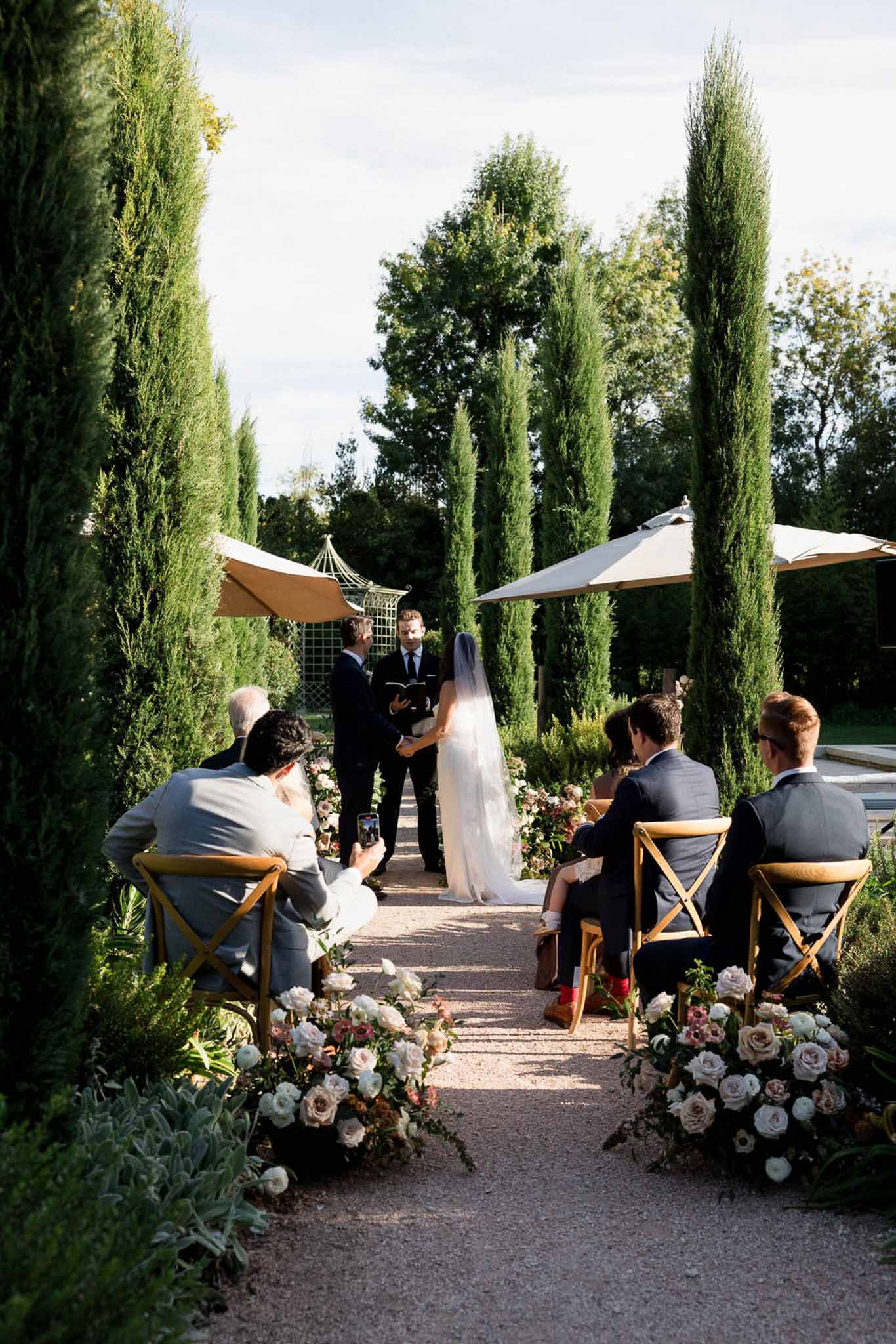 Ceremony with cypress trees, blush rose aisle clusters, and gold cross-back chairs in formal garden