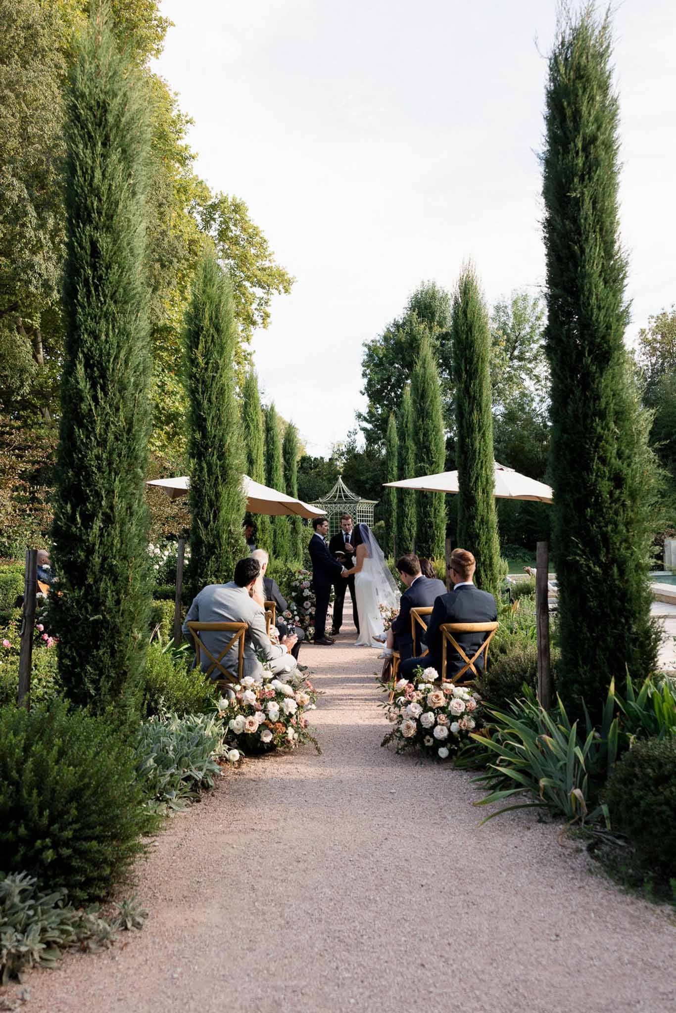 Outdoor garden wedding ceremony with couple at altar flanked by cypress trees and seated guests along flower-lined aisle