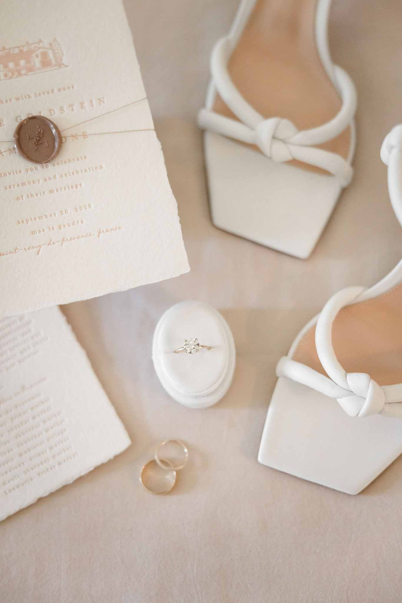 Deckle-edged invitation with wax seal, velvet ring box, gold bands, and white bow-detail bridal heels