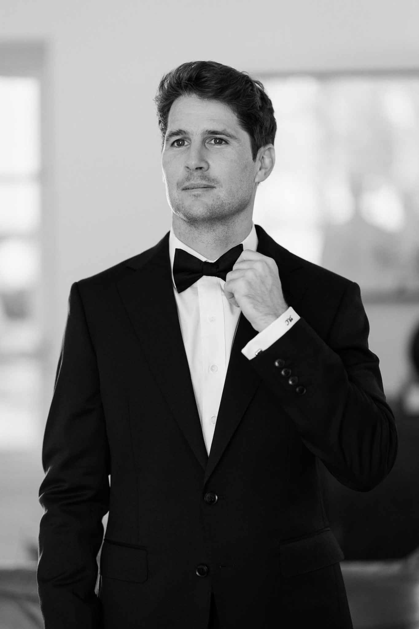 Black and white portrait of groom adjusting bow tie in tuxedo with monogrammed cufflinks during getting ready