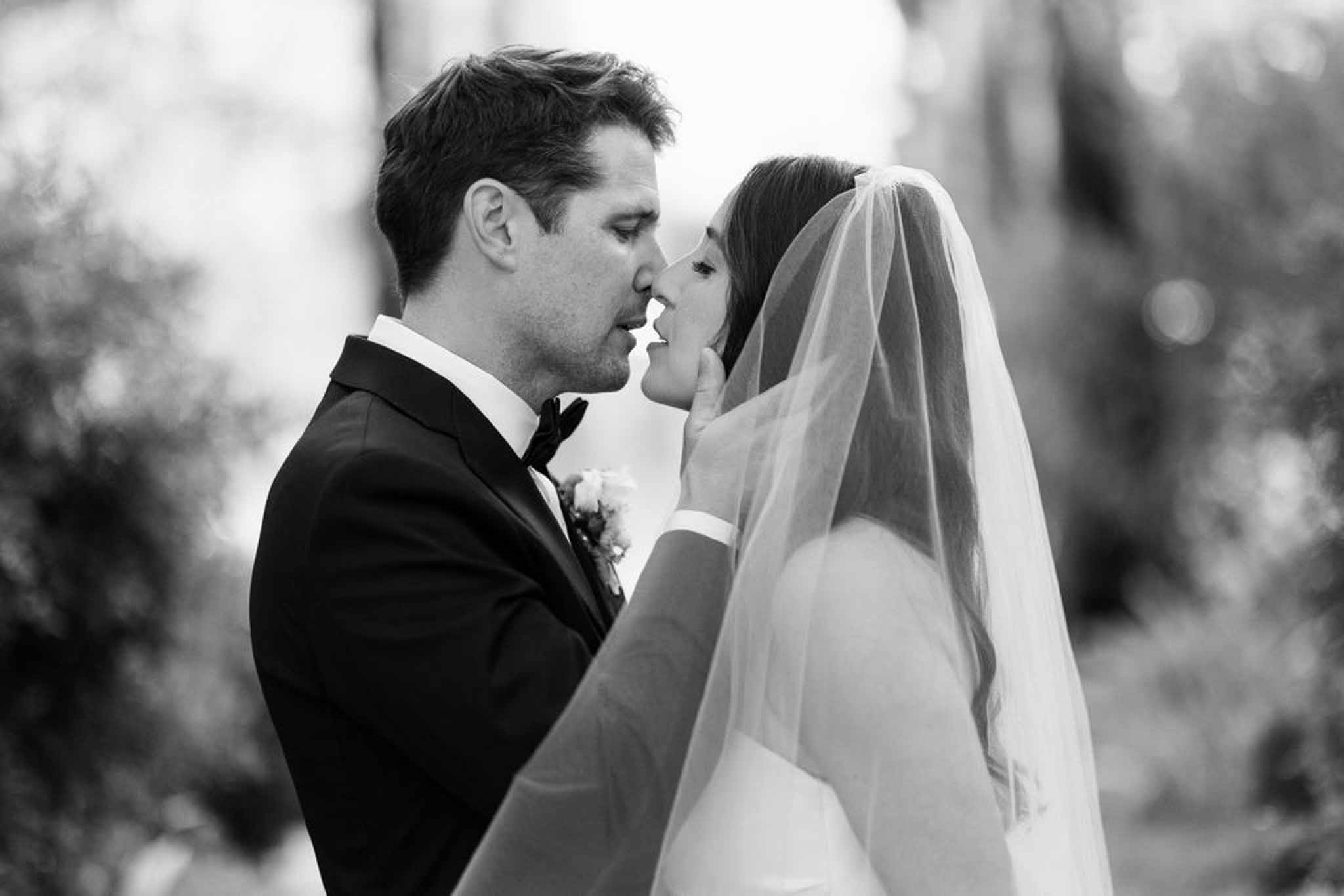 Black and white close-up of couple nearly kissing with flowing veil and blurred foliage background
