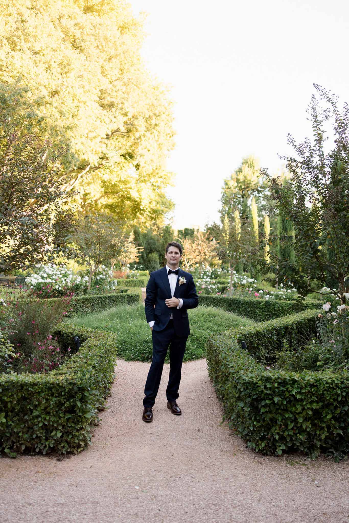 Groom in navy tuxedo standing on gravel path in formal French parterre garden at golden hour