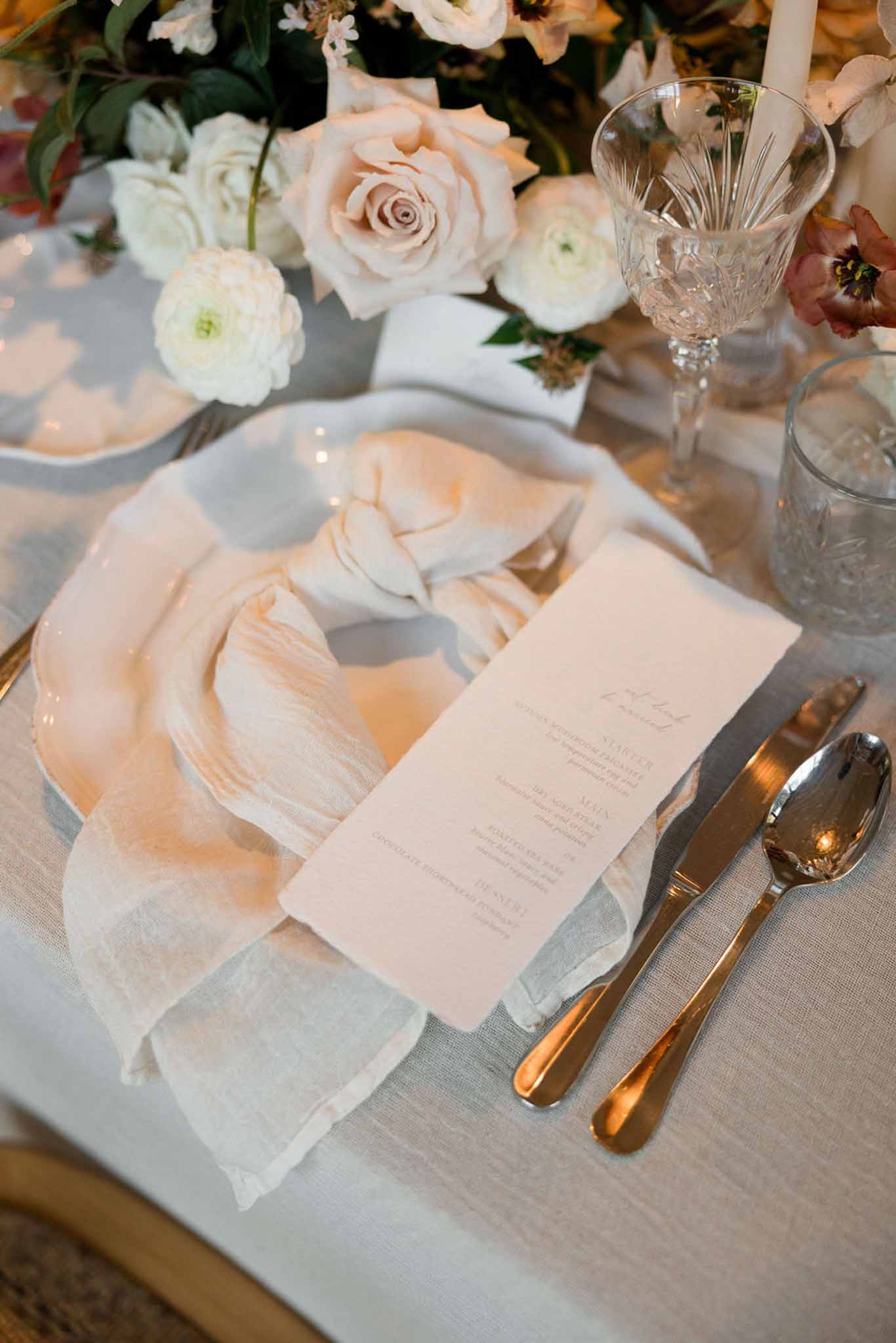 Place setting with grey charger blush menu card gold cutlery and centerpiece of blush roses burgundy anemones on blue linen