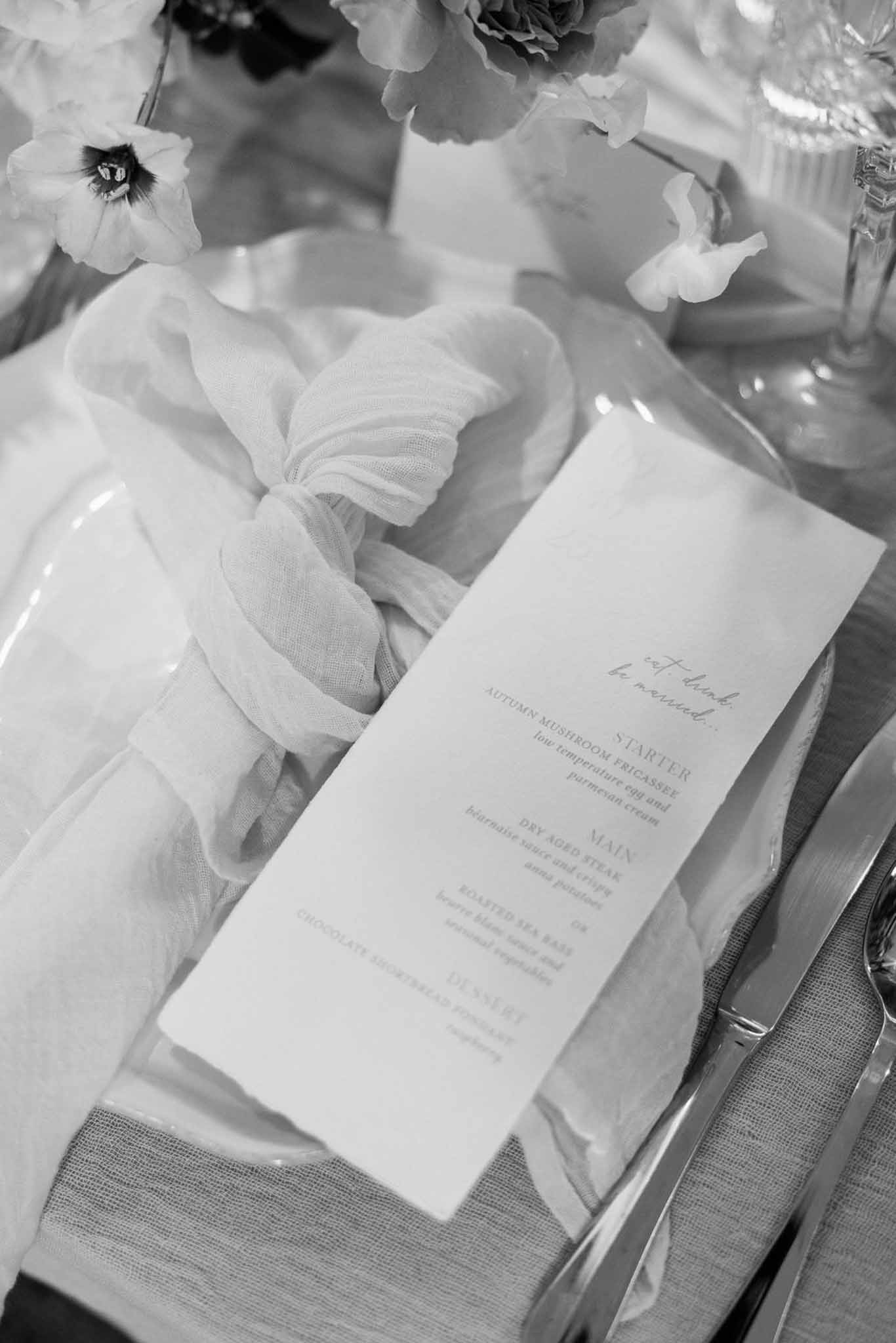 Black and white close-up of printed menu card on gauze napkin with silver cutlery and anemone stems