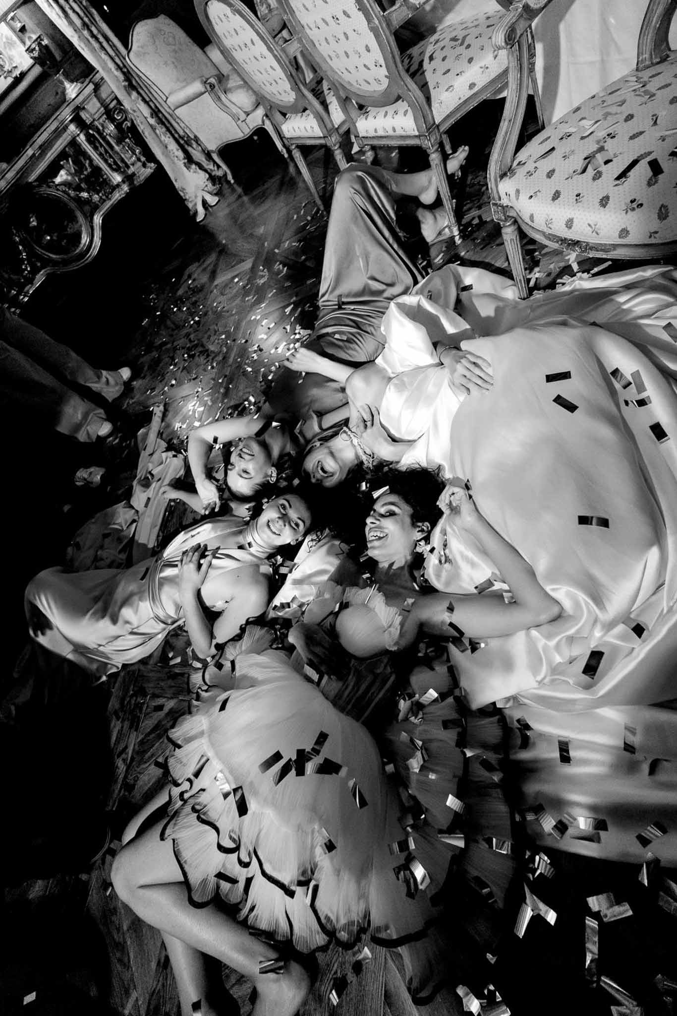 Overhead black-and-white shot of bride and bridesmaids lying on floor laughing surrounded by confetti in chateau ballroom