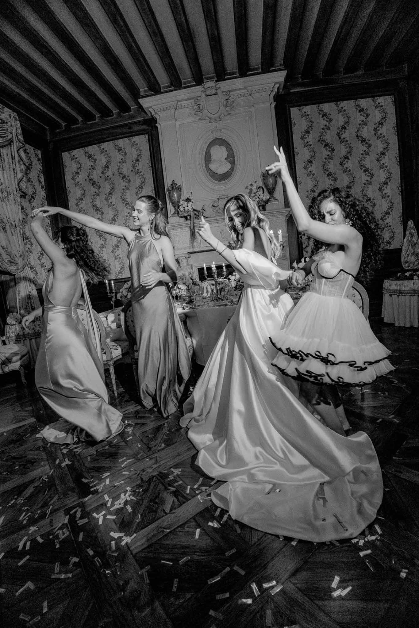 Black and white four women dancing on confetti-strewn parquet in coffered-ceiling chateau ballroom