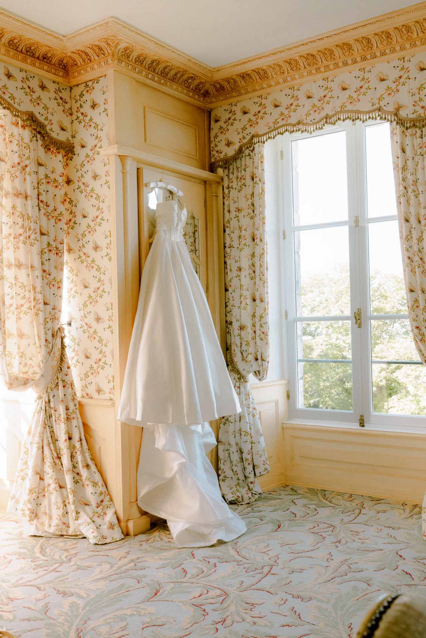 White strapless bridal gown hung on carved armoire in ornate chateau room with floral wallpaper and gilded molding