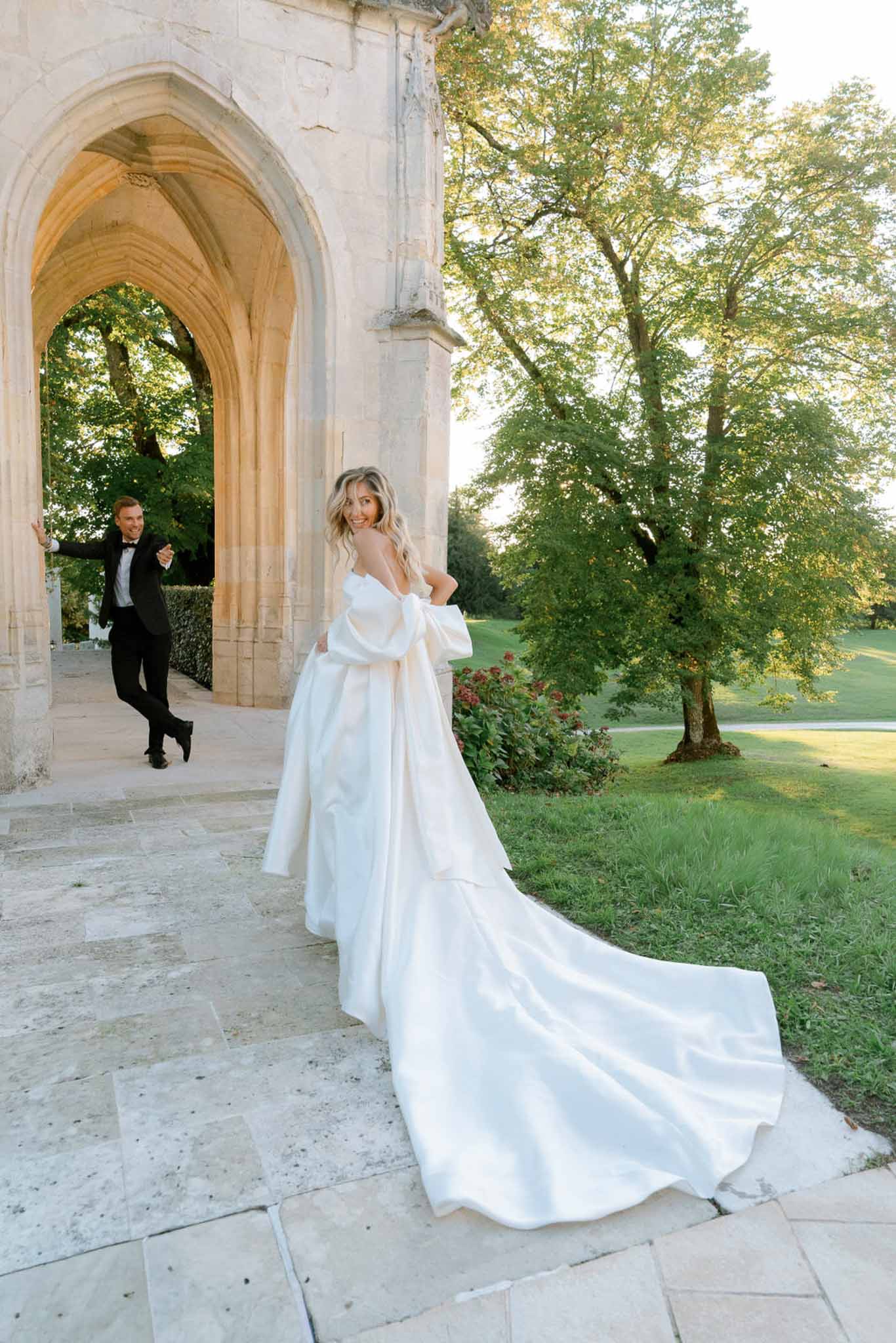 Bride in satin ball gown with cathedral train looking back as groom walks through Gothic stone arch