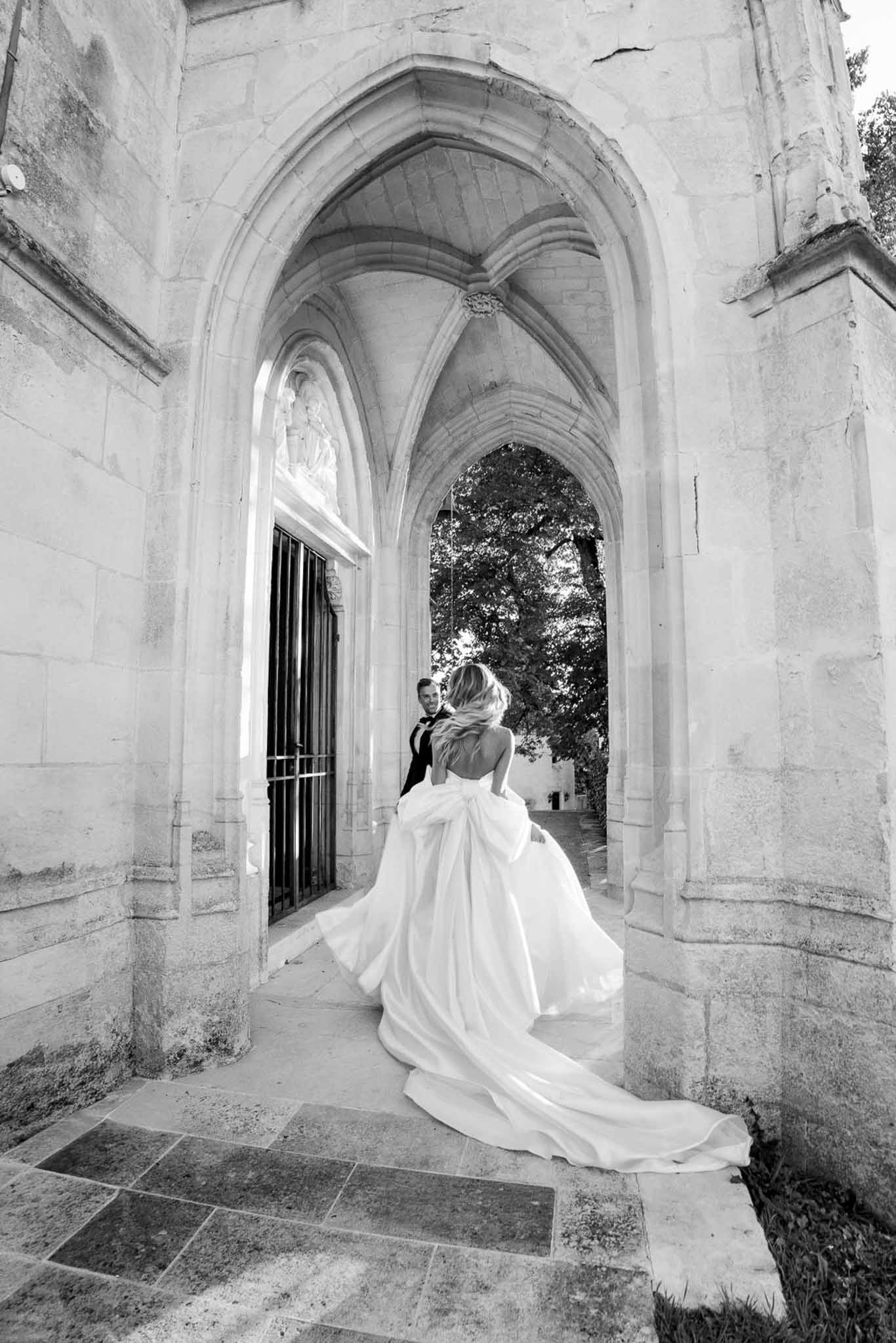 Black and white portrait of bride in bow-back ballgown walking through Gothic stone arches toward groom