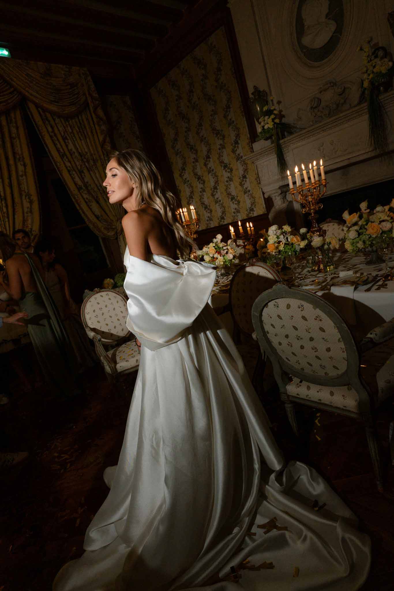 Bride in bow-back satin gown at candlelit table with ivory roses and gold candelabras in chateau salon