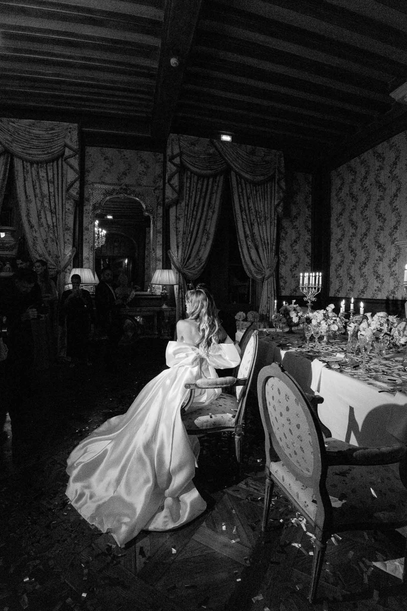 Black and white rear view of bride in dramatic bow-back gown seated at candlelit chateau head table