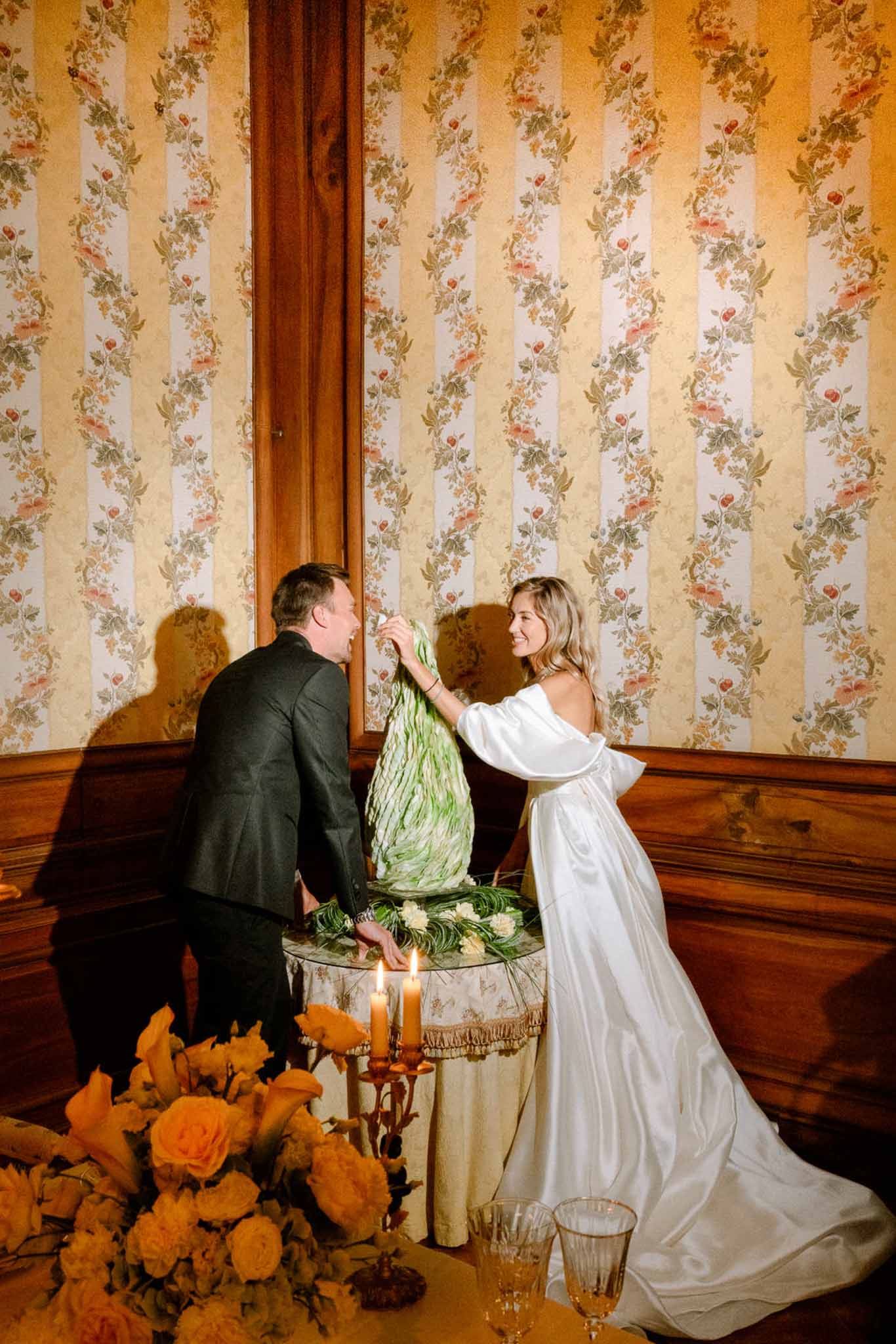 Couple with sculptural green leaf cone beside orange roses, amber candles, and vintage floral wallpaper