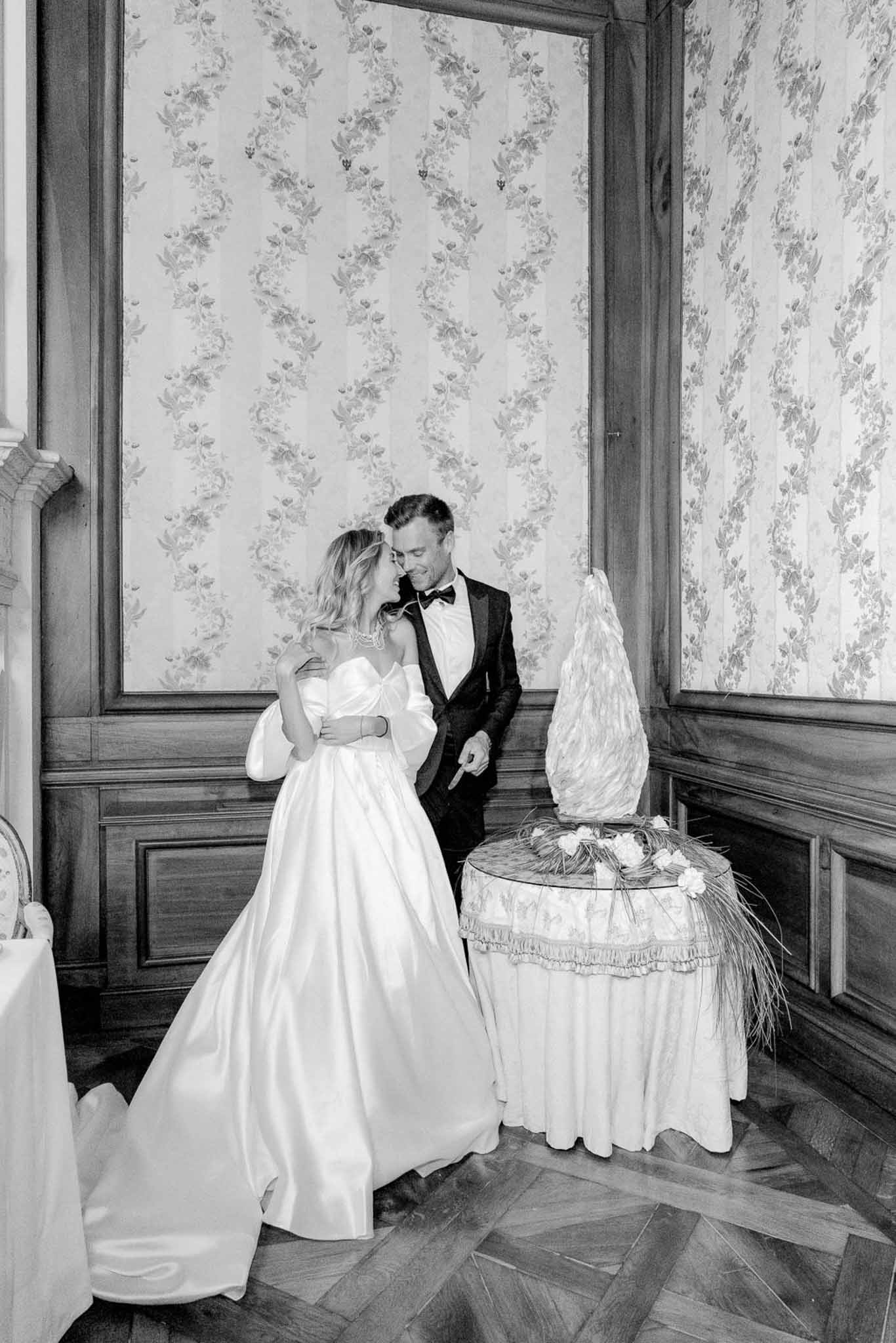 Black-and-white image of bride and groom cutting a sculptural ruffled wedding cake in a chateau ballroom