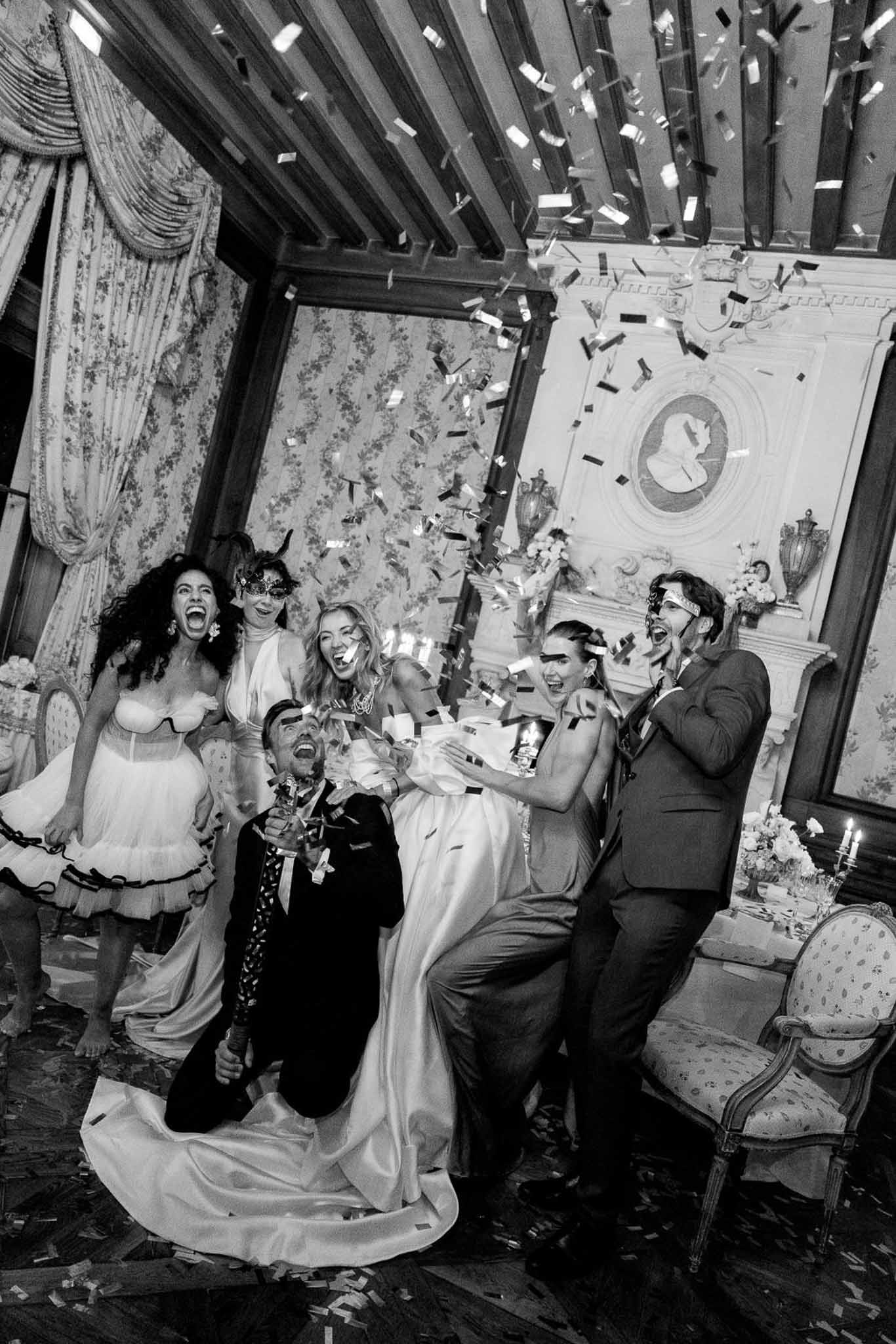 Black-and-white photo of bride and groom with guests in masquerade masks as confetti falls in ornate chateau ballroom
