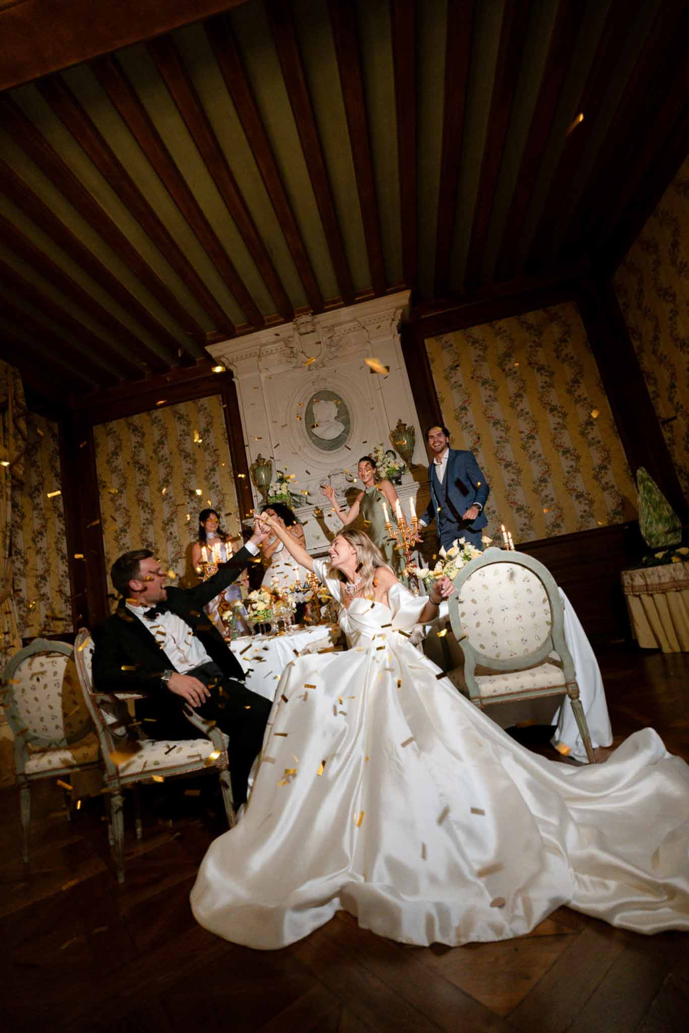 Bride and groom celebrate with gold confetti at chateau reception table with candelabras and ornate fireplace backdrop