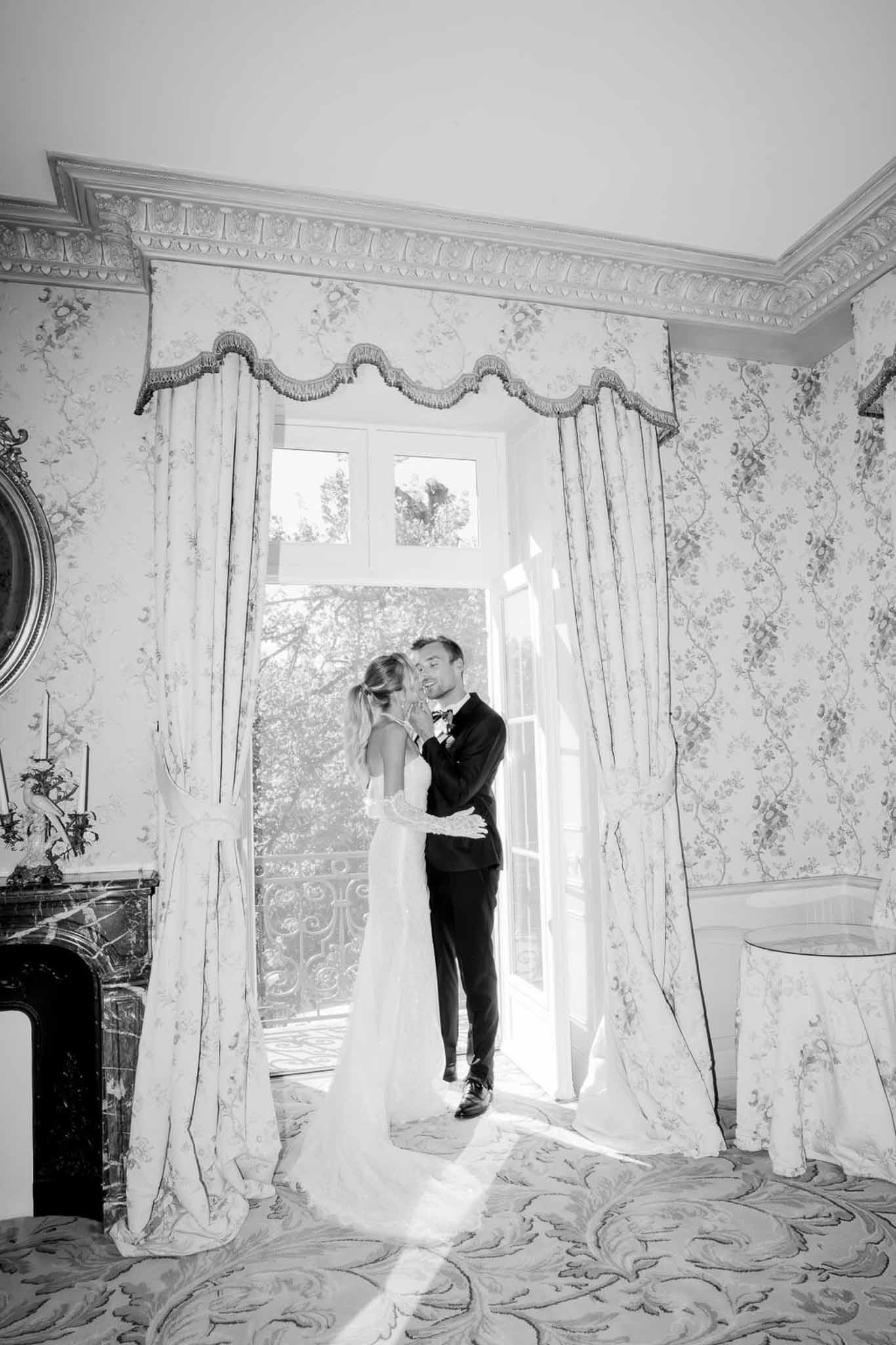 Bride and groom silhouetted before backlit French doors in ornate chateau salon in B&W