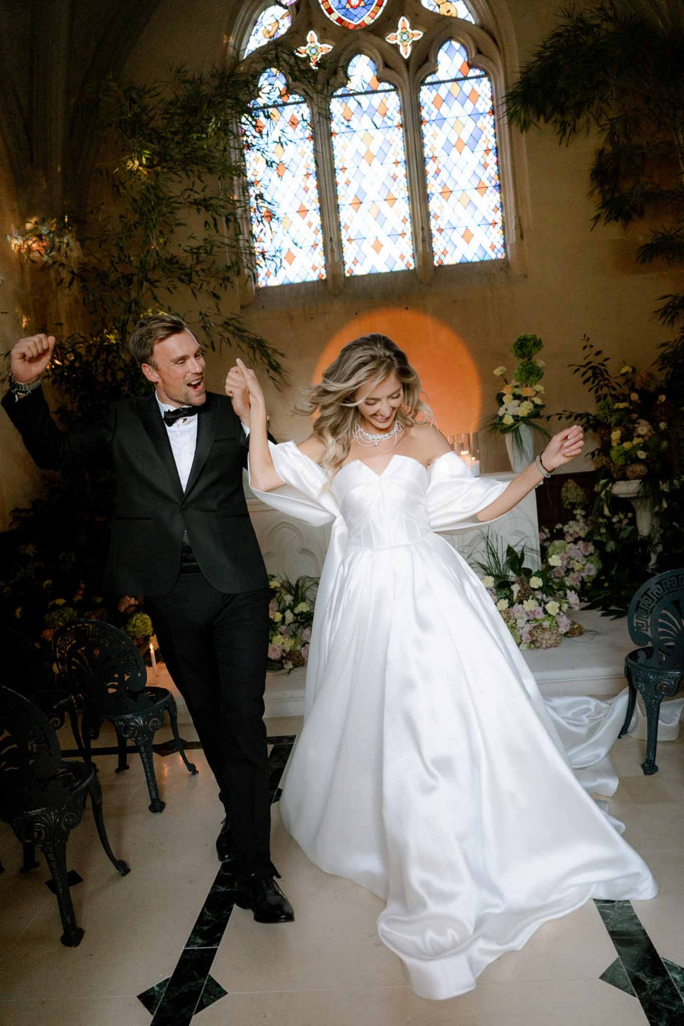Bride in puff-sleeve ballgown and groom dancing in chapel with stained glass windows and greenery altar