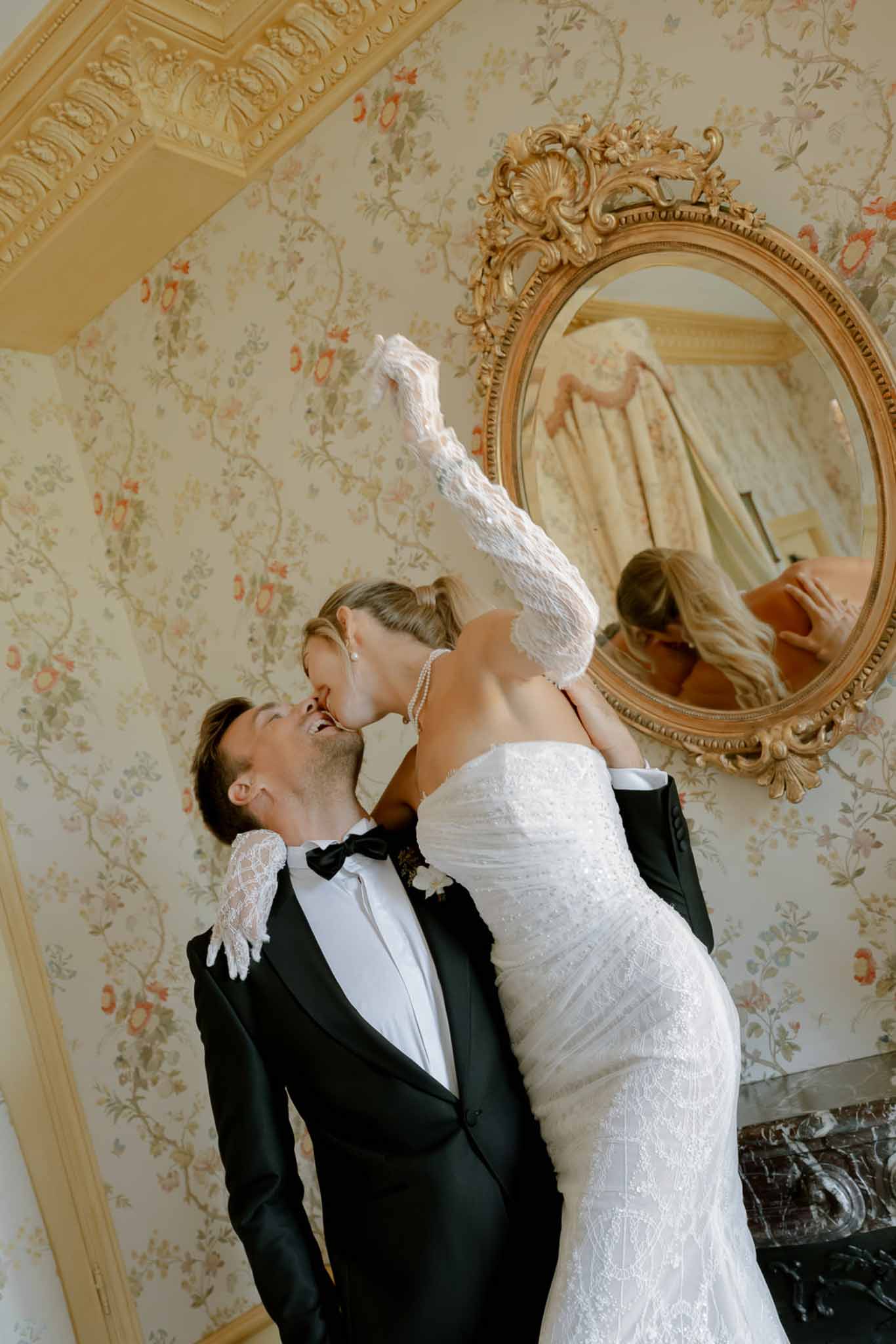 Groom lifting bride in a kiss inside ornate chateau room with gilt mirror and marble fireplace