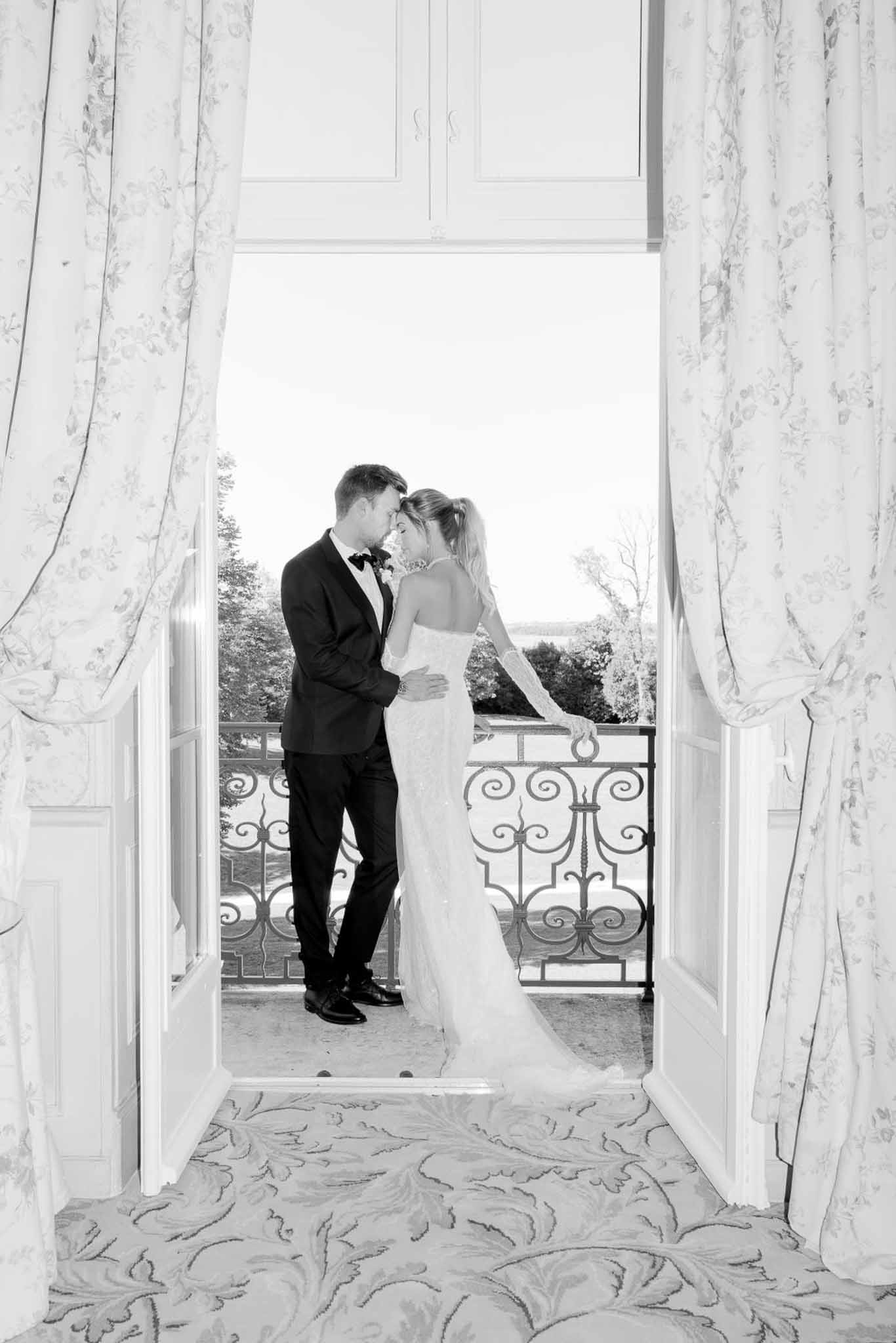 Black and white portrait of bride and groom with foreheads touching on chateau balcony framed by French doors