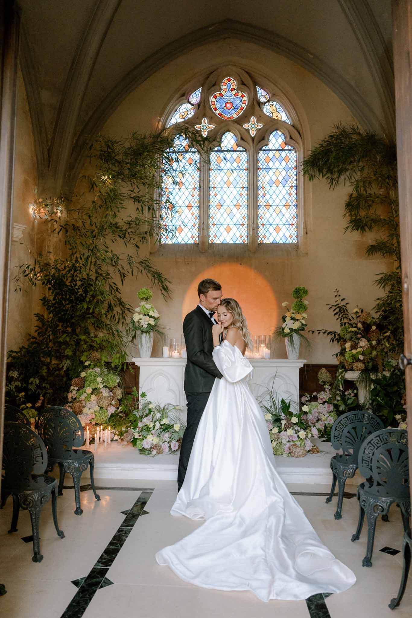Couple embracing at chapel altar with Gothic stained glass, trailing greenery, and white floral display