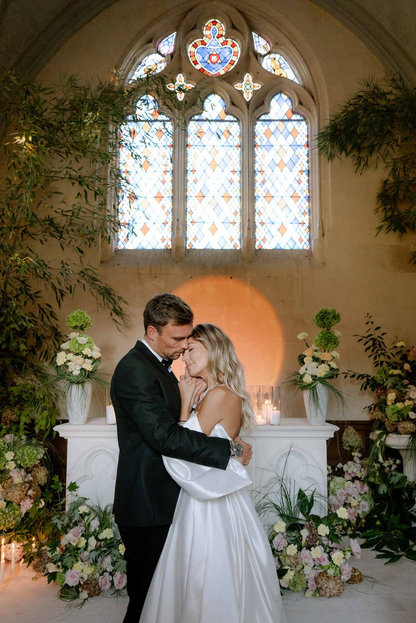 Couple embracing at altar with green hydrangea and blush roses beneath Gothic stained-glass window