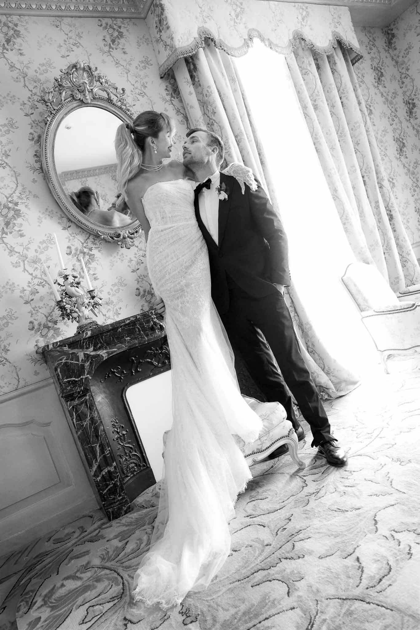 Bride and groom in near-kiss pose in ornate chateau room with floral wallpaper and marble fireplace in B&W