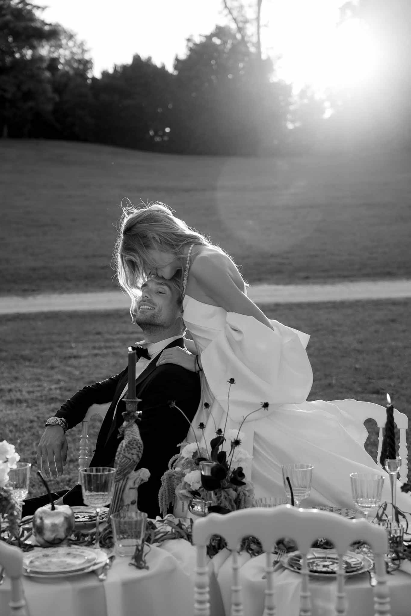 Black and white bride kissing groom cheek at styled sweetheart table with backlit sun flare