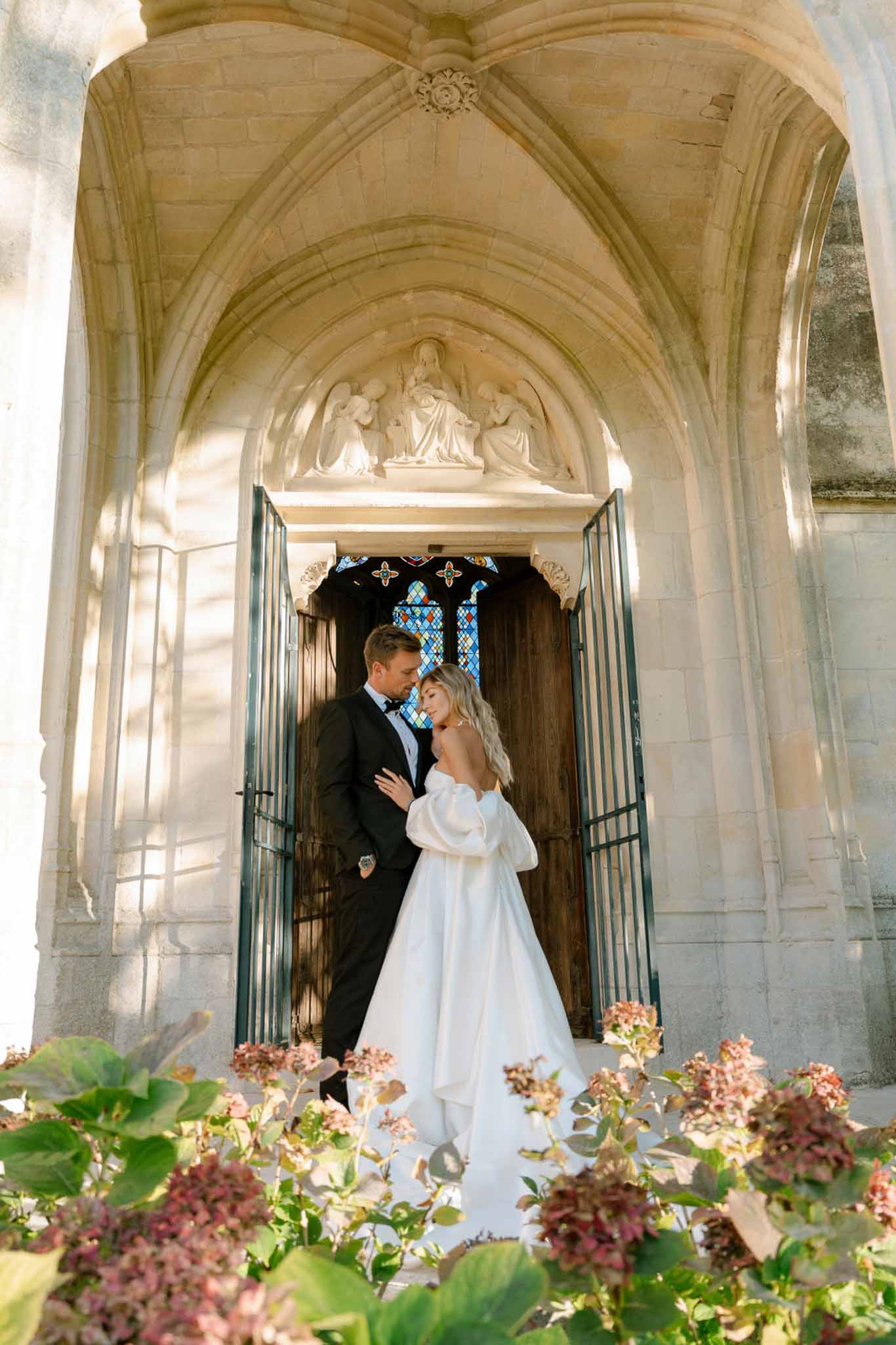 Bride and groom embracing in Gothic chapel doorway framed by hydrangeas with carved stone arch above