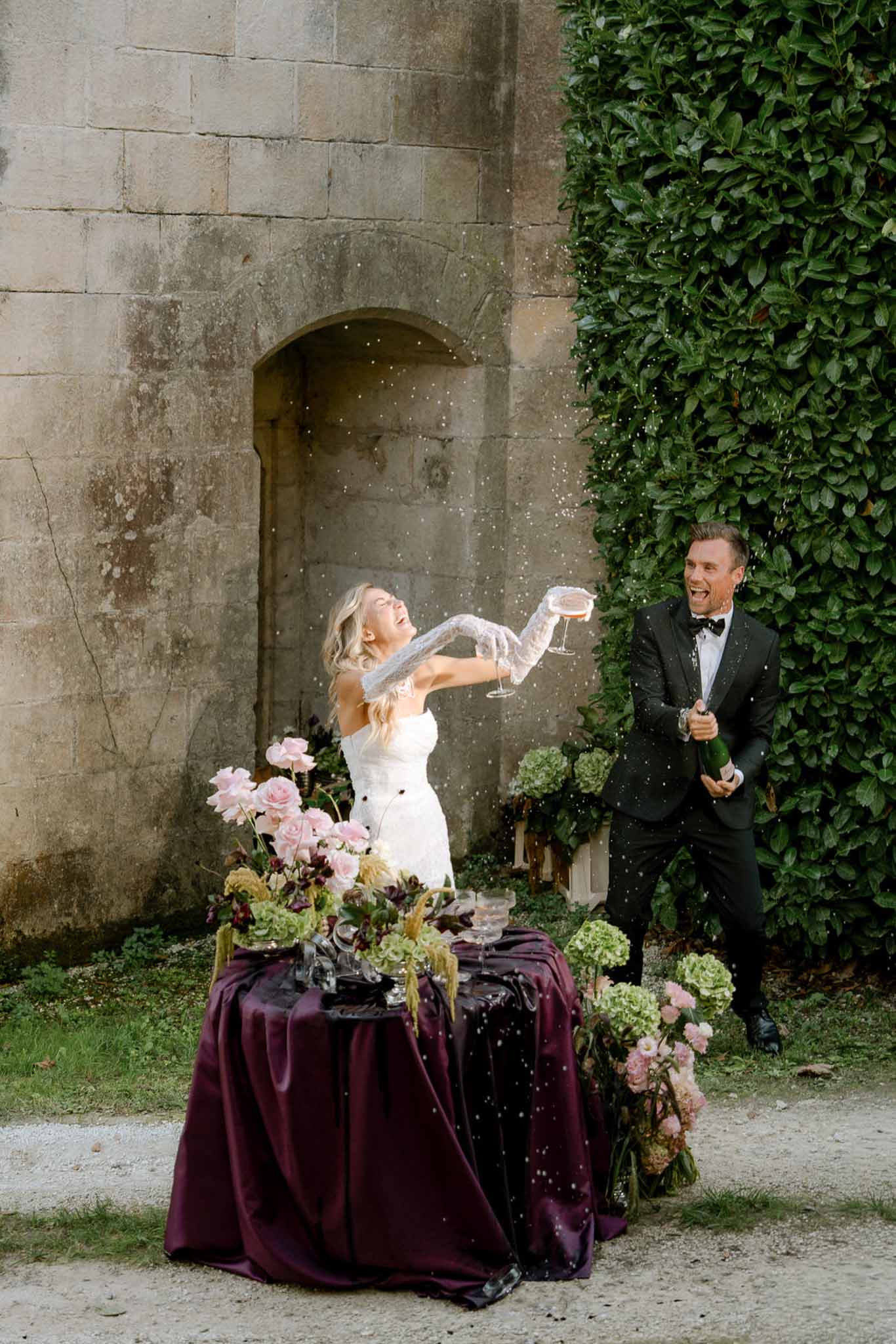 Groom popping champagne as bride laughs beside burgundy satin table with blush and pink rose arrangement on gravel