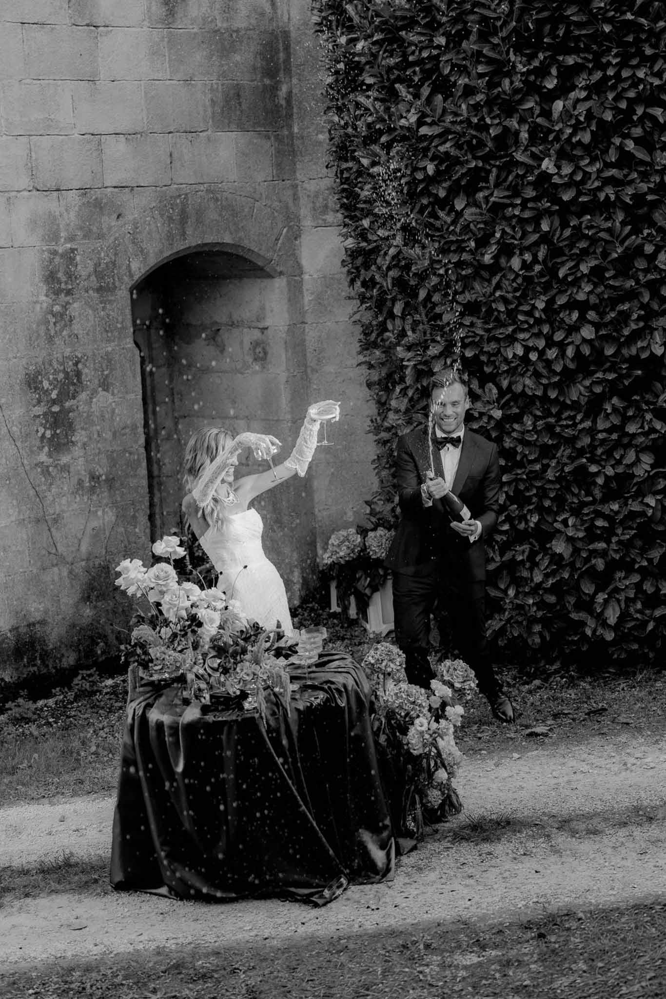 Black-and-white image of bride and groom spraying champagne bottle outside chateau with rose and hydrangea arrangement