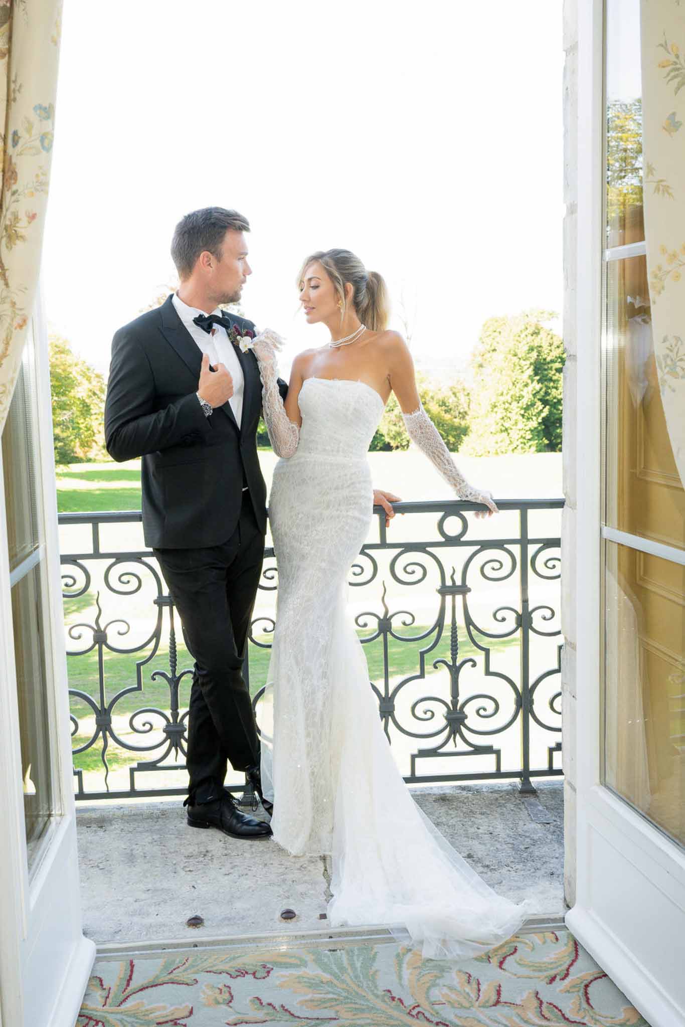 Couple on wrought iron balcony bride in beaded gown with pearl choker groom in tuxedo with burgundy boutonniere