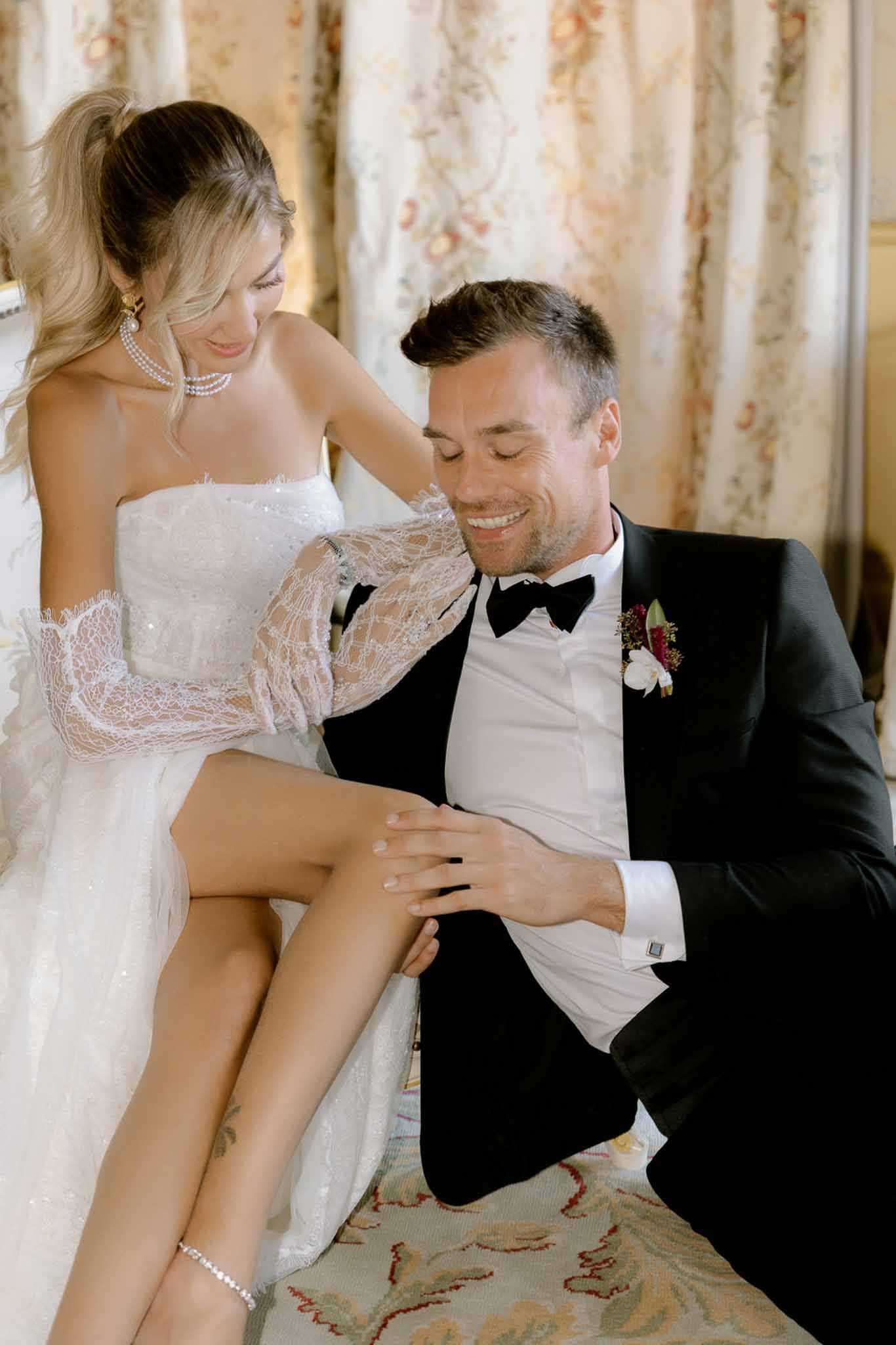 Groom kneels beside seated bride in pearl necklace and beaded gown before floral curtains in chateau